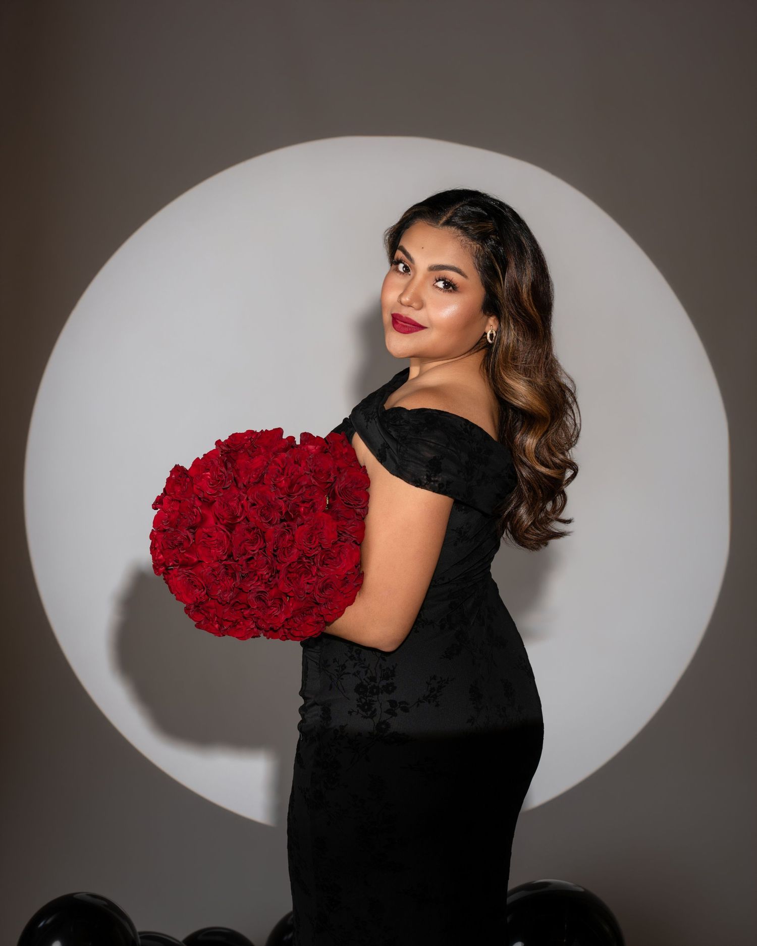 Woman in a black fitted dress and red lipstick poses sideways holding a large red rose bouquet before a circular backdrop.