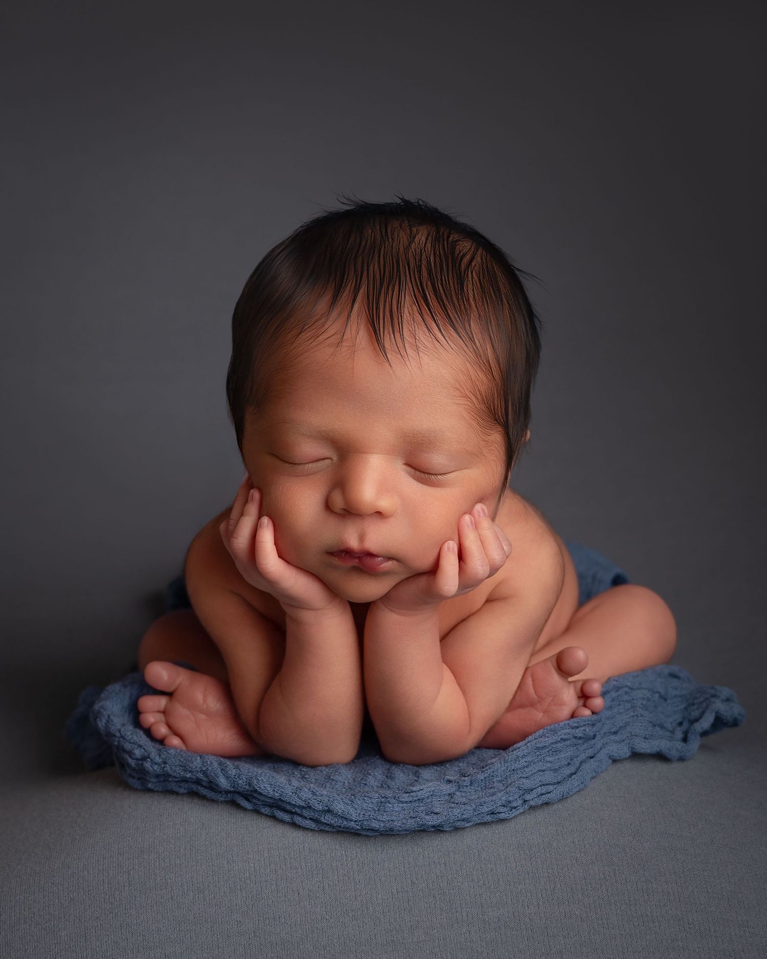 Peaceful sleeping newborn baby posed in froggy position on blue wrap on gray backdrop.
