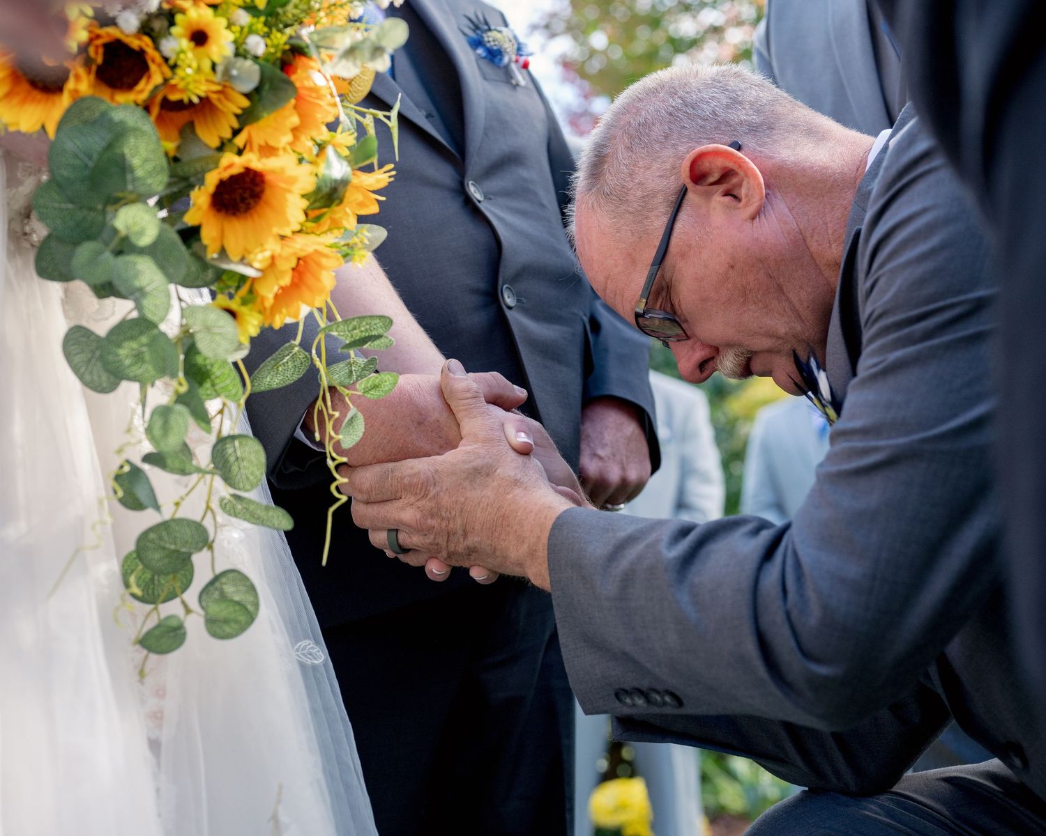 Emotional wedding moment as a man bows his head in prayer while holding the bride and grooms hands.