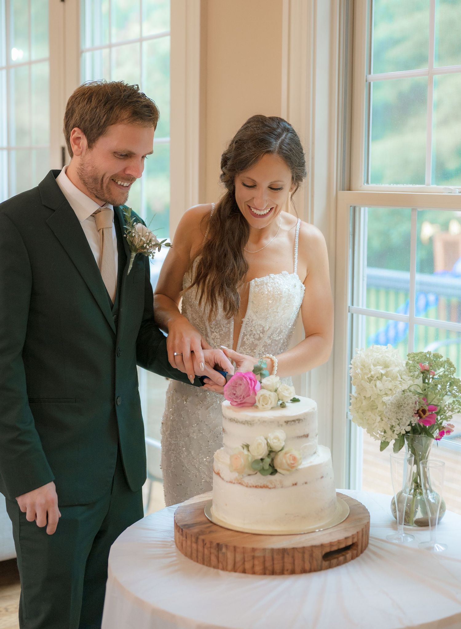 Bride and groom joyfully cut their two-tier semi-naked wedding cake adorned with fresh pink and white flowers.