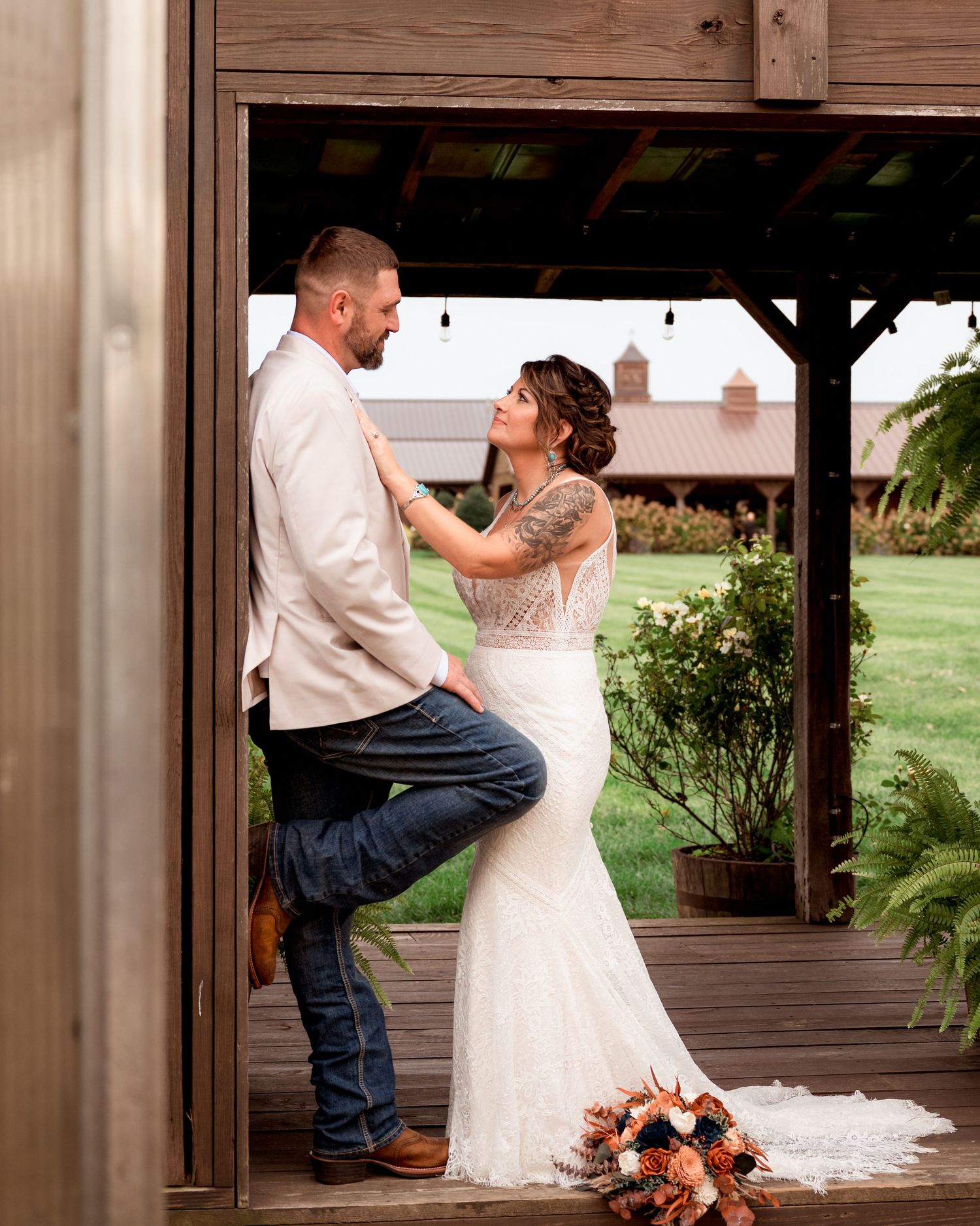 Bride and groom share a tender moment in a rustic barn doorway, with a colorful bouquet at their feet.