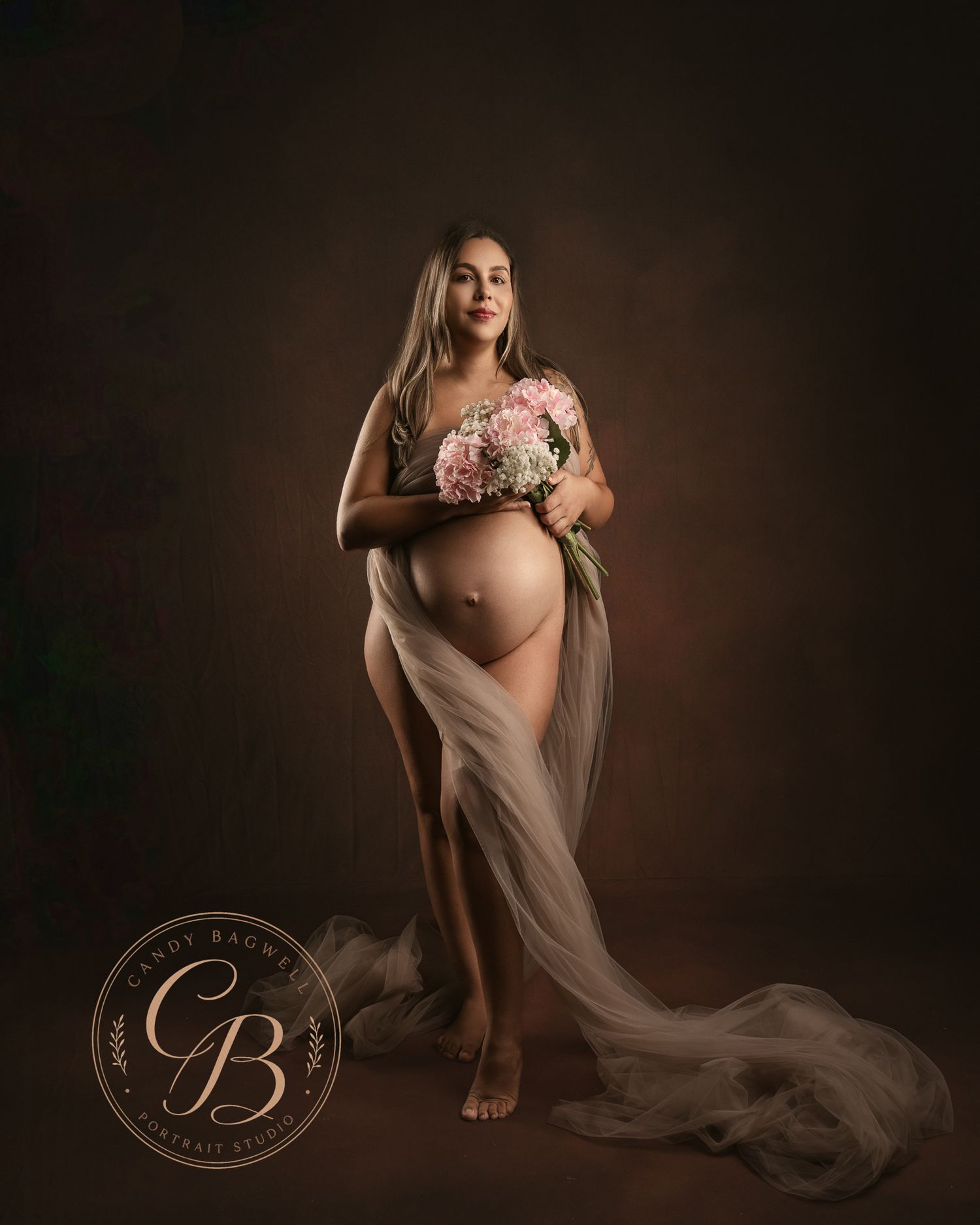 Pregnant woman posing with pink flowers, draped in sheer fabric, studio maternity portrait by Cindy Brown Photography.