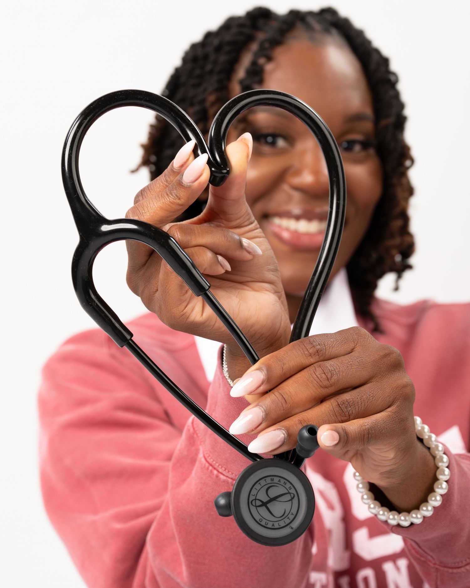 Smiling nurse holding black stethoscope in heart shape toward camera with pearl bracelet.