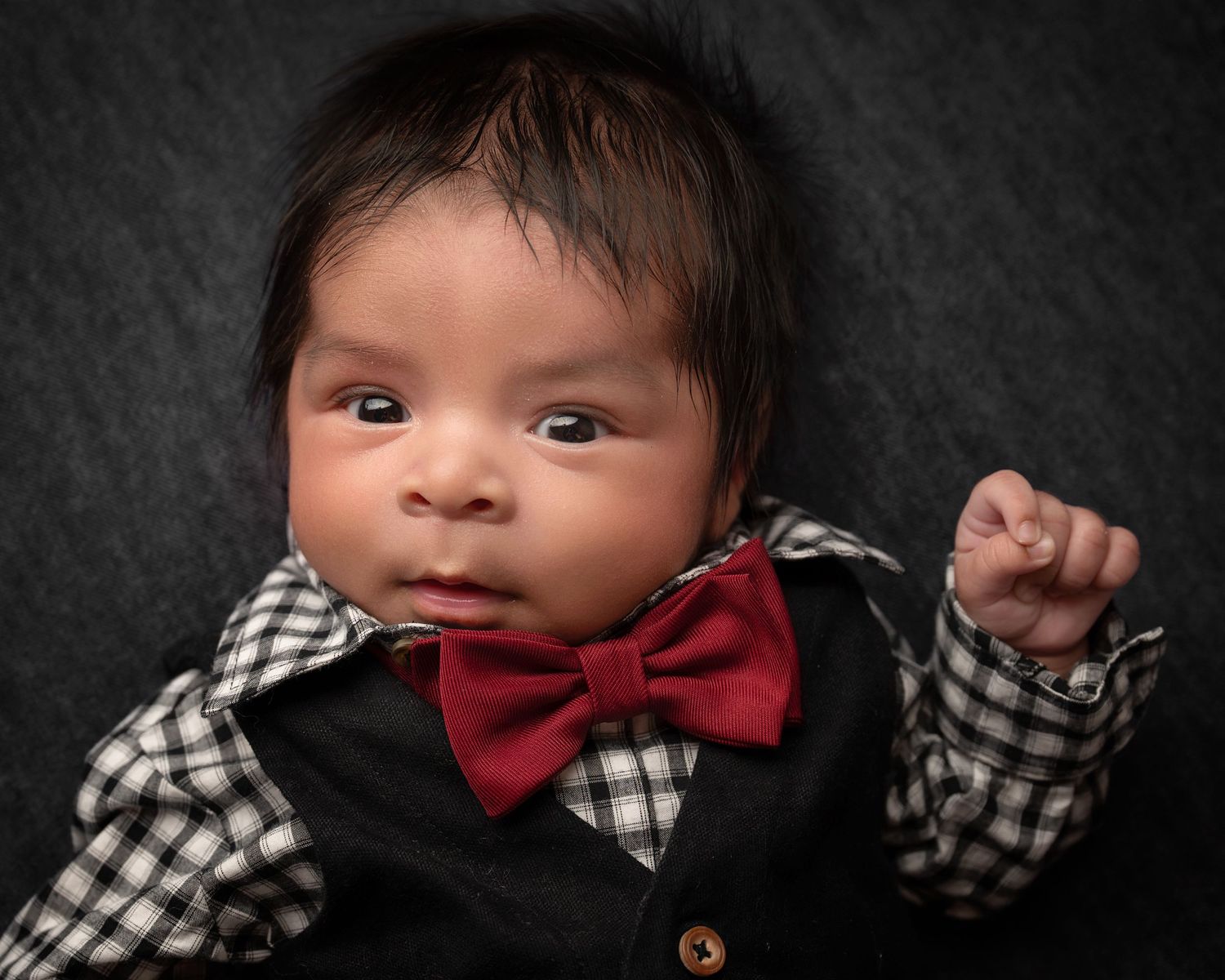 Alert newborn baby boy in black tuxedo outfit with red bow tie on dark background.