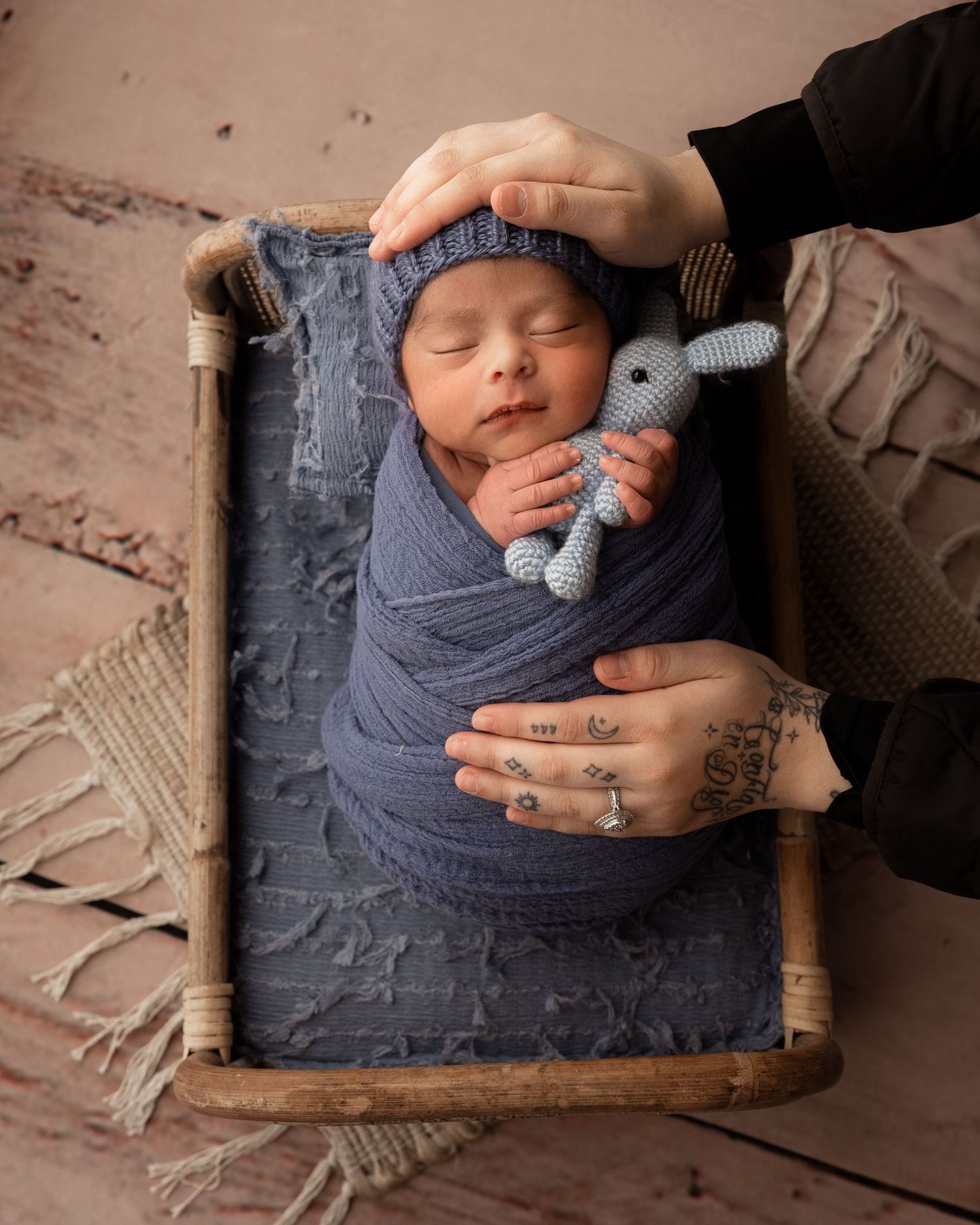 Sleeping newborn in blue swaddle and knit hat held by tattooed parent hands in wooden bed.