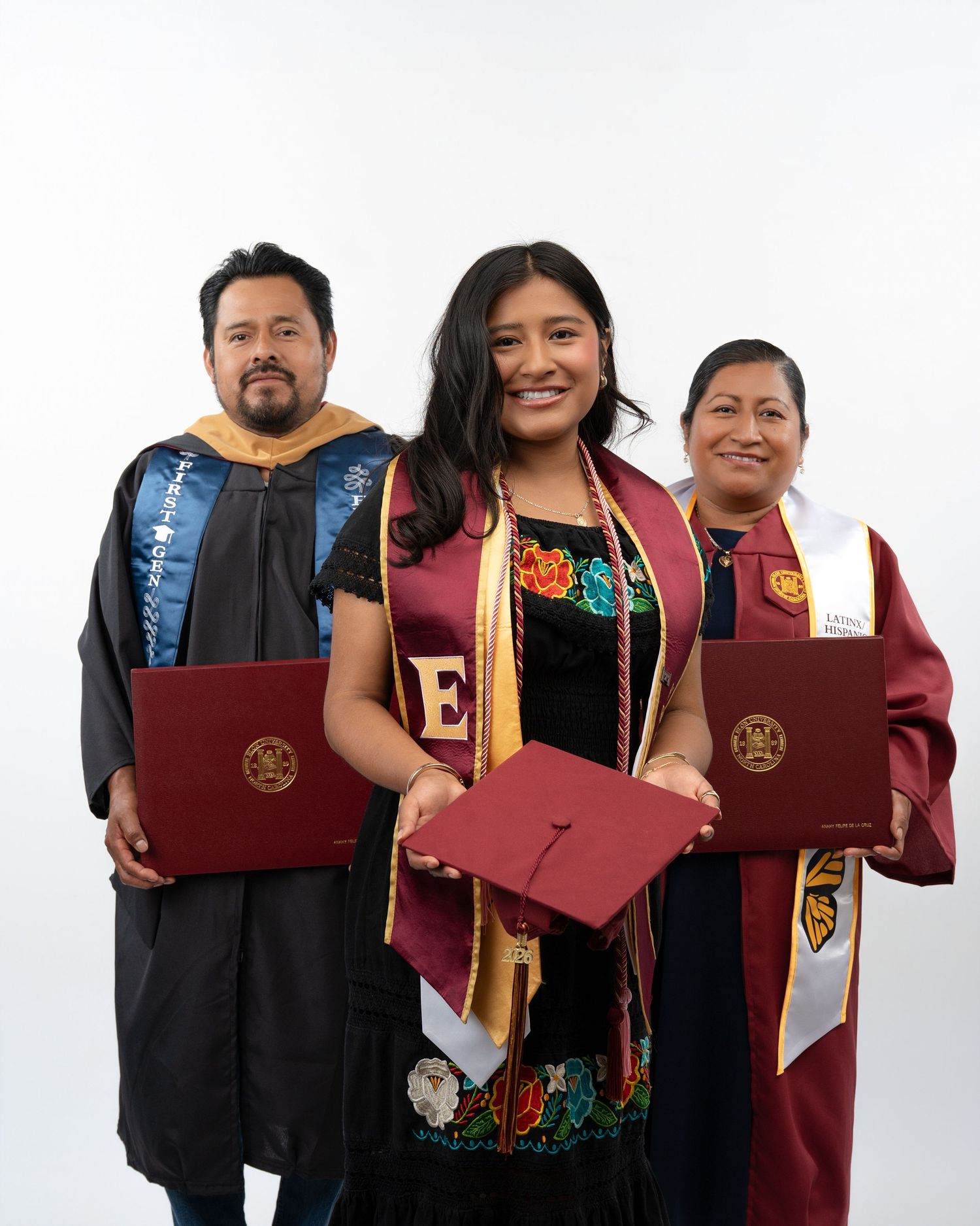 Three graduates in academic regalia proudly display red diploma covers in family graduation portrait.