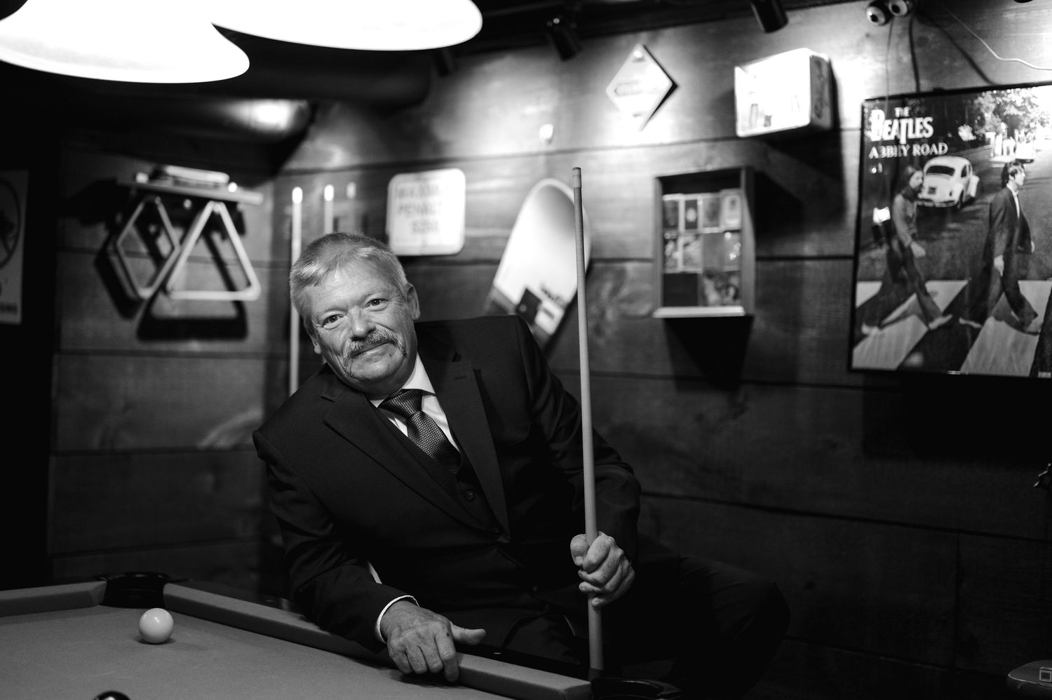 Elegant man in suit posing with pool cue at billiard table in black and white portrait photo.