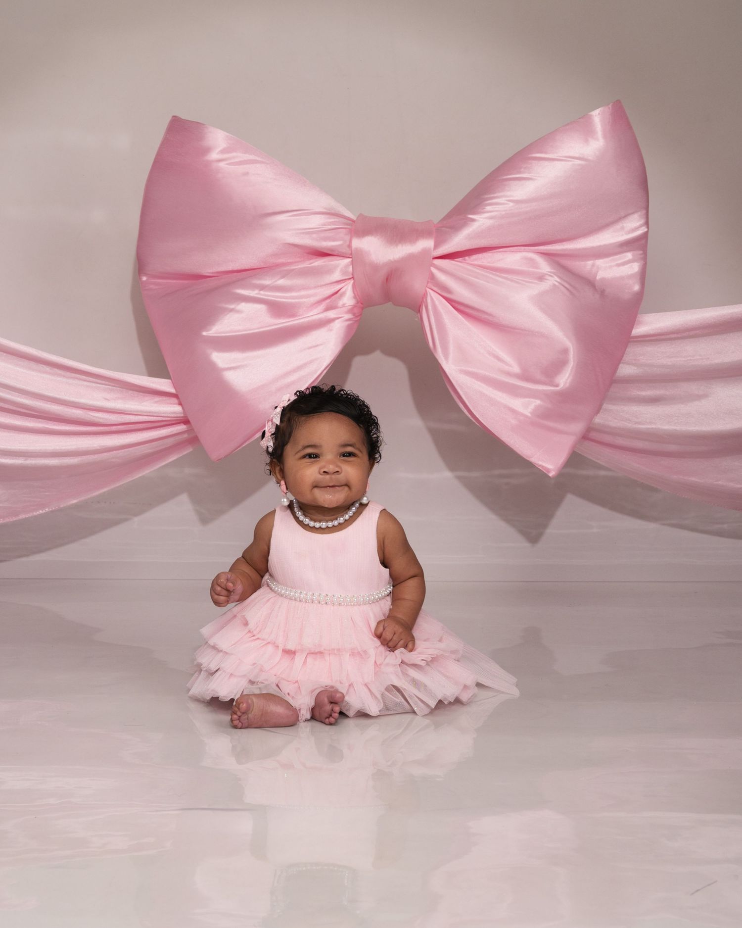 Adorable baby girl in pink tutu dress smiles in front of large decorative pink satin bow backdrop.