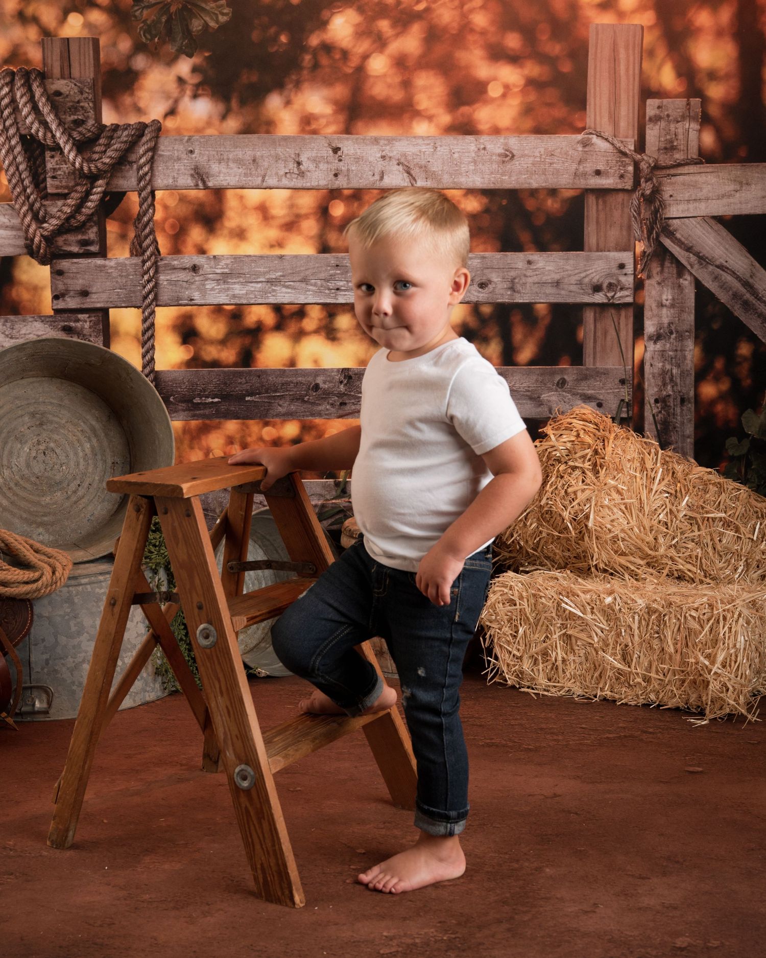 Toddler boy in white shirt and jeans posing by wooden ladder in farm-themed studio.