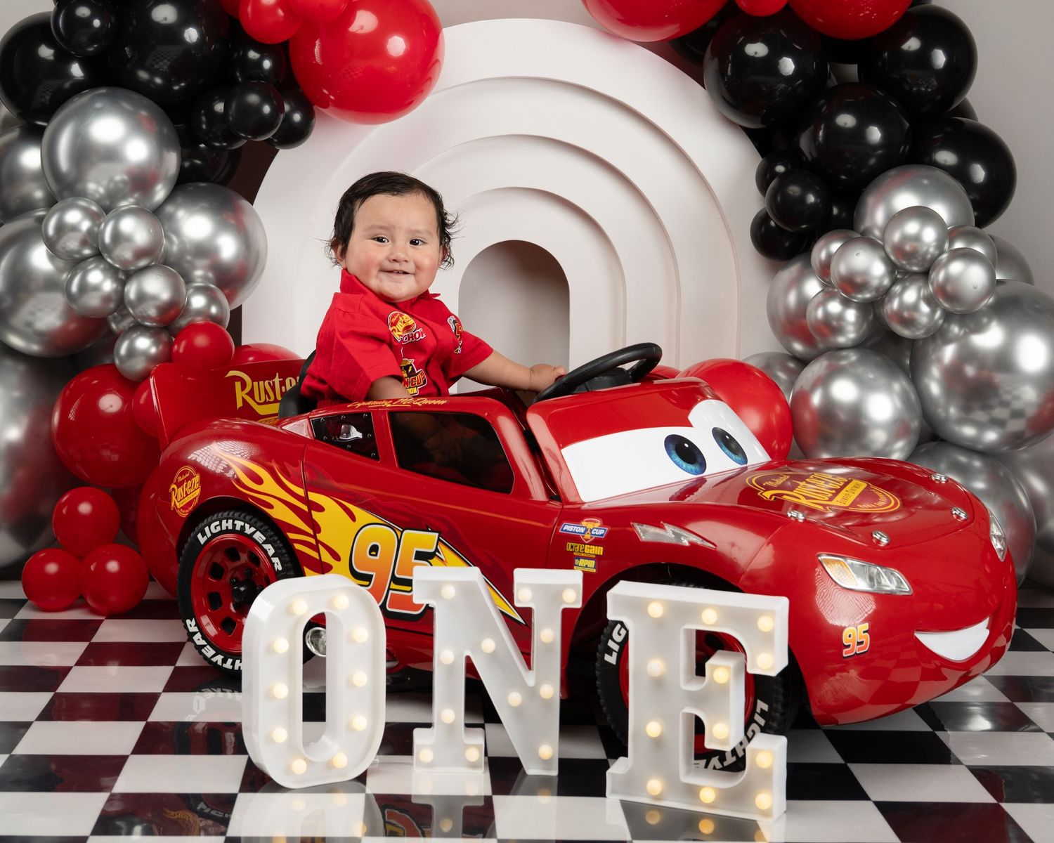 A happy baby boy sits in a Lightning McQueen ride-on car surrounded by a black and silver balloon arch.