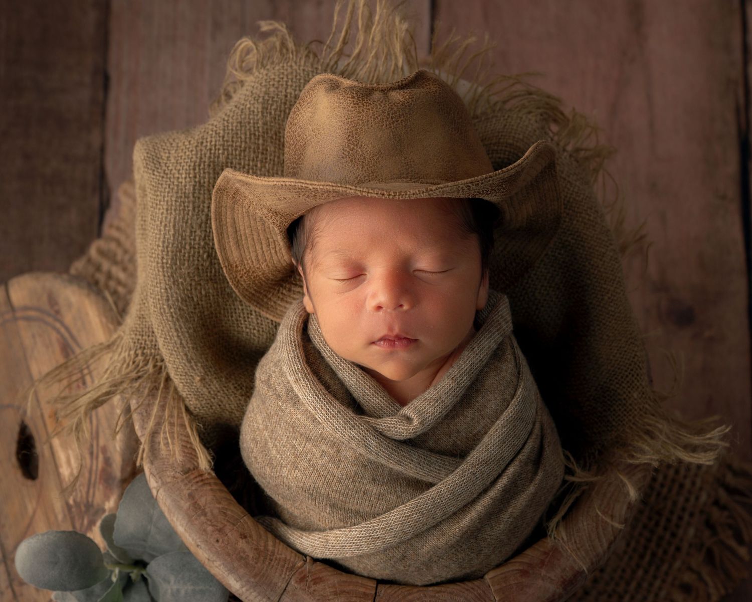 Sleeping newborn wrapped in burlap swaddle wearing tiny cowboy hat in rustic wooden spool prop.