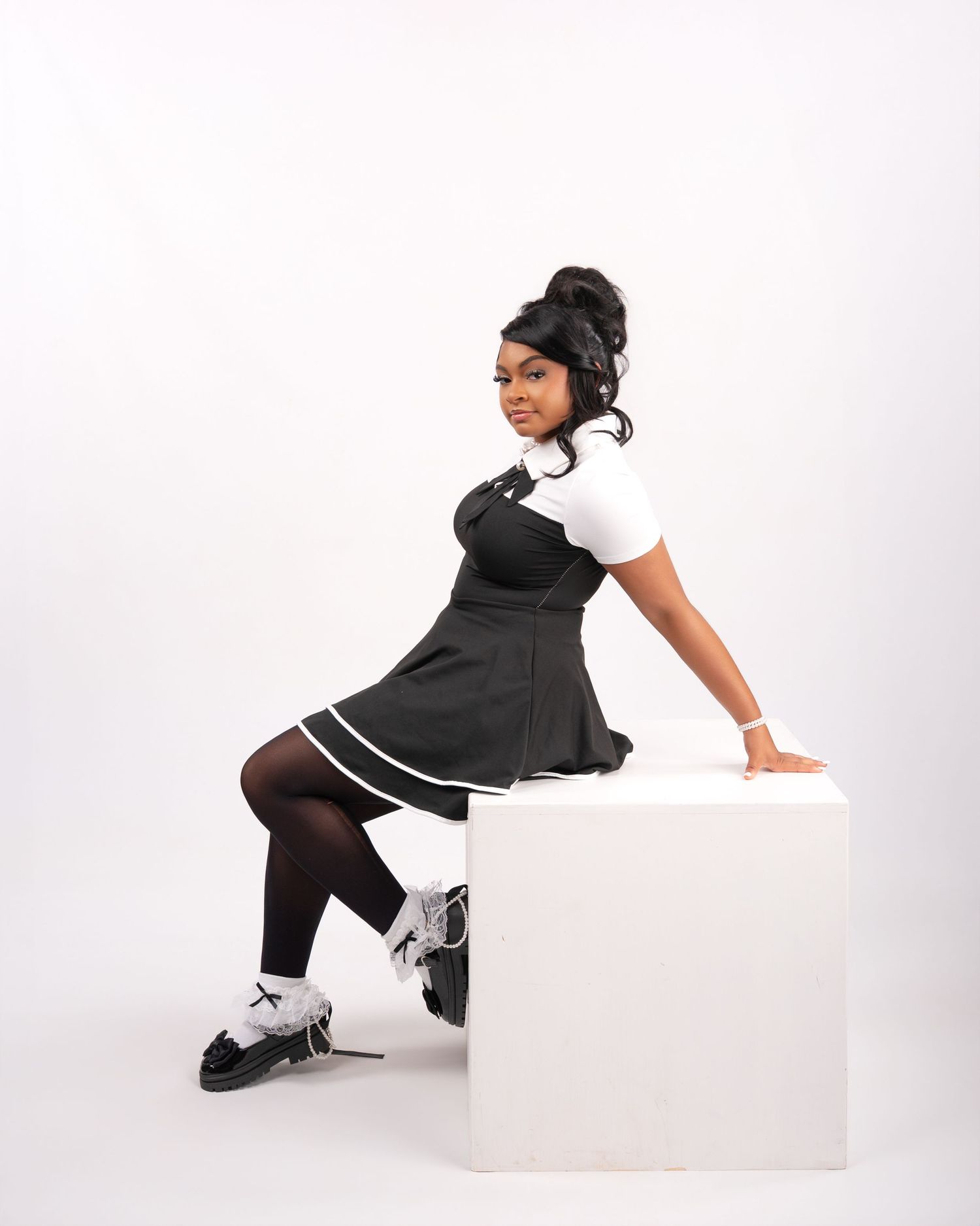 Young woman in black pinafore dress with white trim, black tights, and platform shoes poses on white cube.