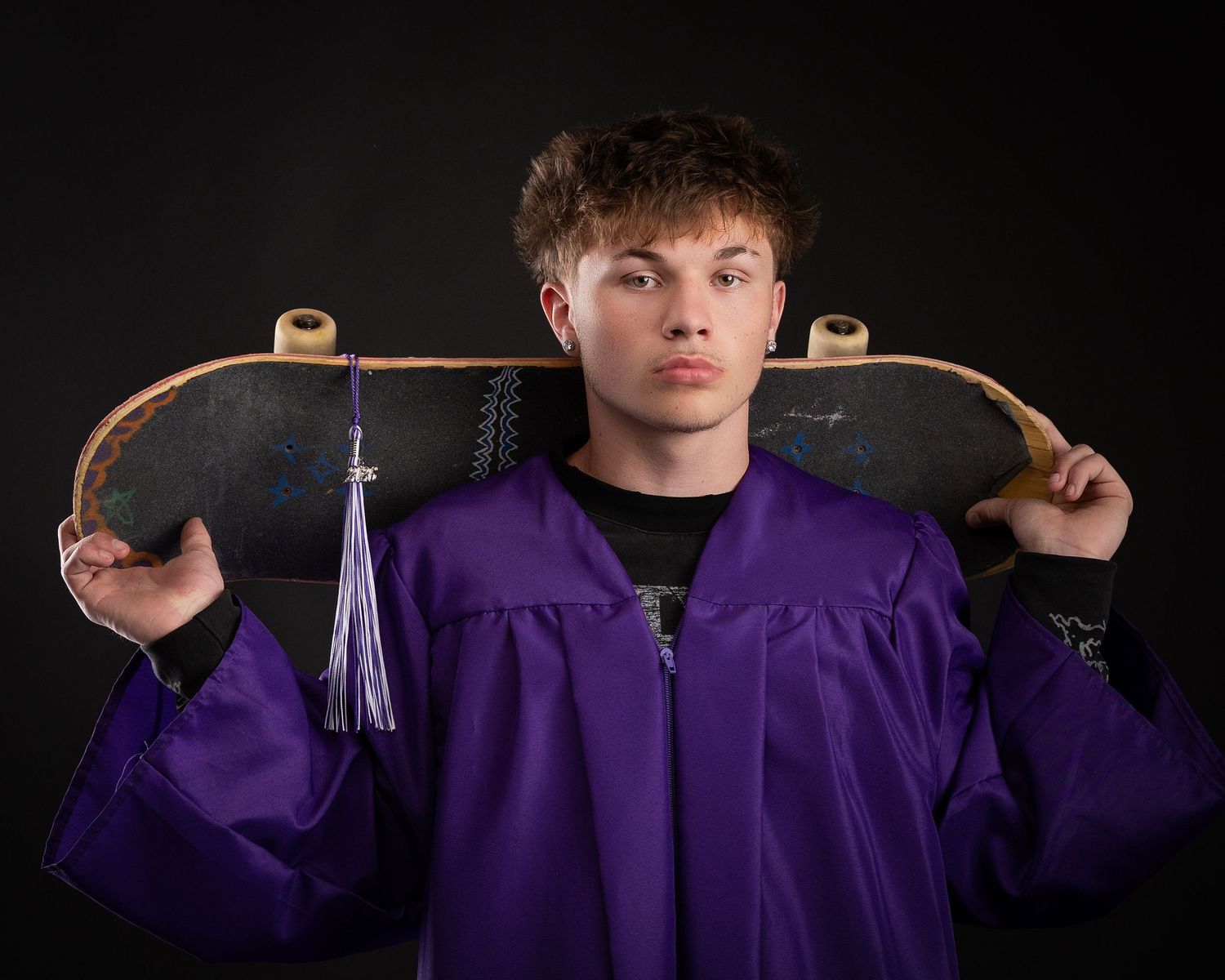 Teen male graduate in purple gown holds skateboard across shoulders against dark background.