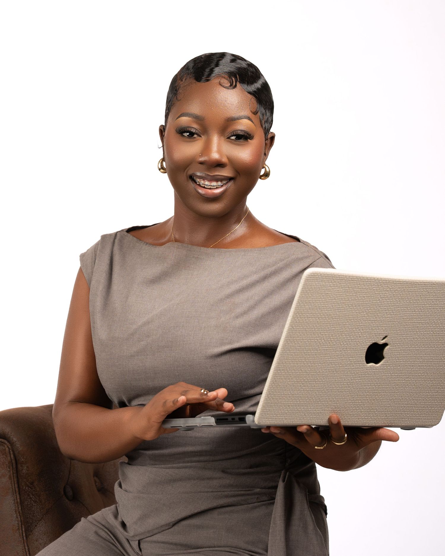 Smiling professional woman in gray dress holding Apple MacBook laptop, seated against white background.