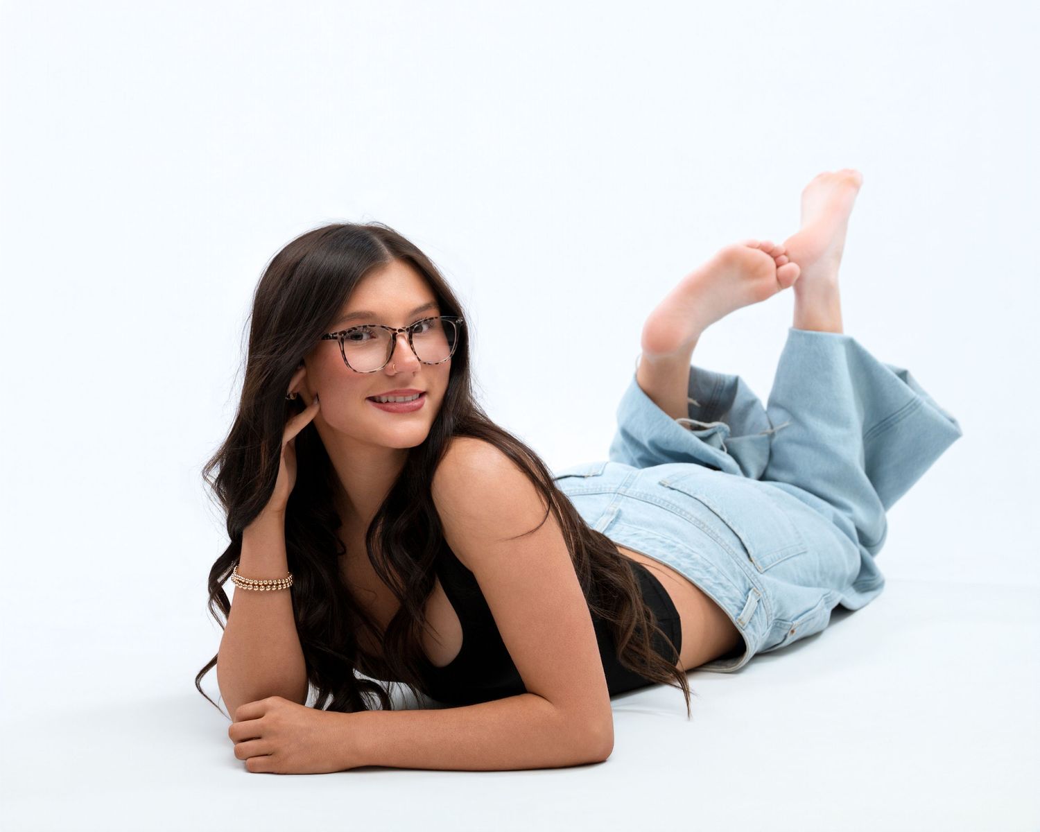 Smiling young woman with long dark hair and glasses lies on her stomach in light blue jeans against a white studio background.