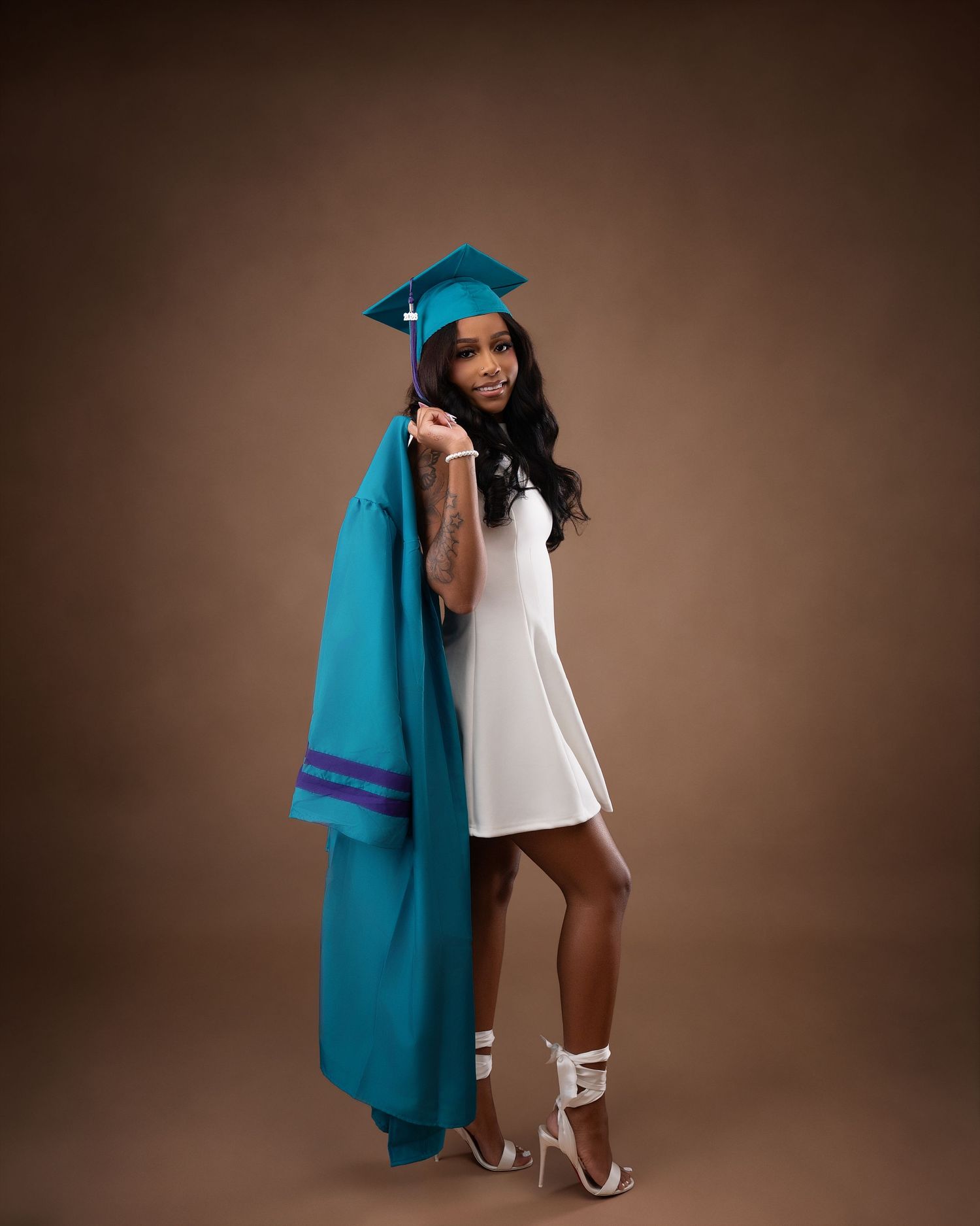 Graduate in teal cap and gown poses in white dress with heels against a warm brown background.