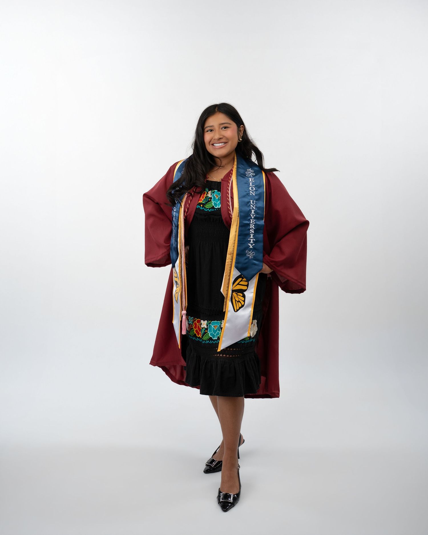 Smiling female graduate in maroon gown with colorful embroidered sash poses in bright studio portrait.