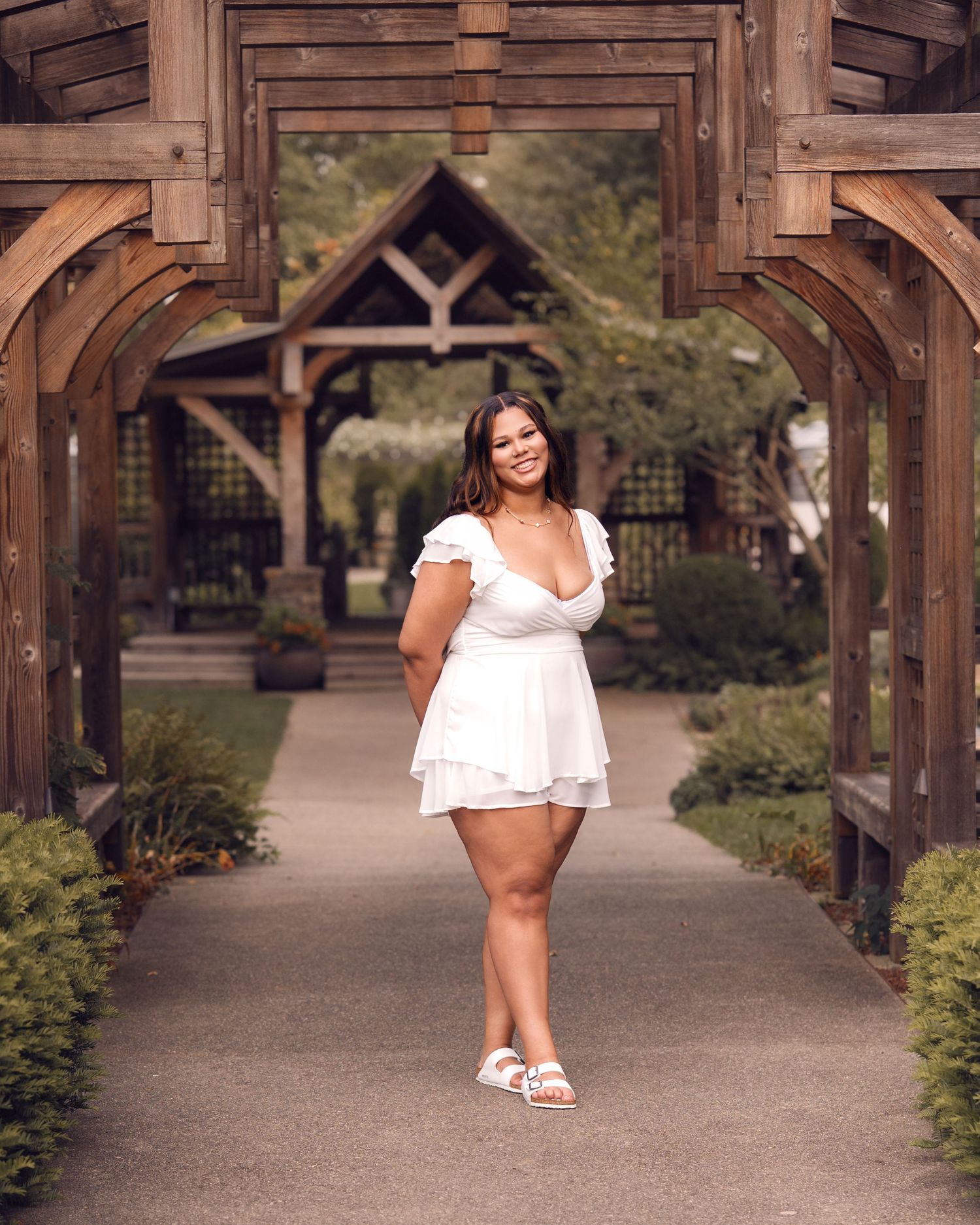 Smiling young woman in white ruffle dress poses under wooden arch in garden setting.