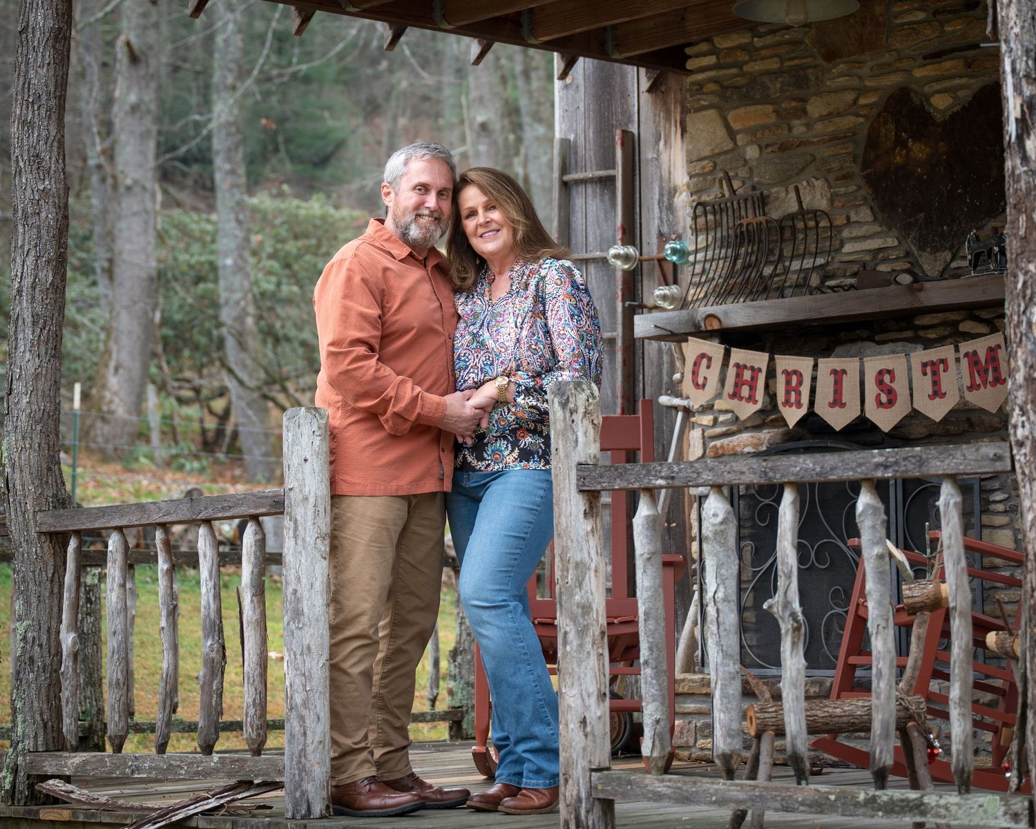 A couple smiles together on a rustic log cabin porch with a Christmas banner.