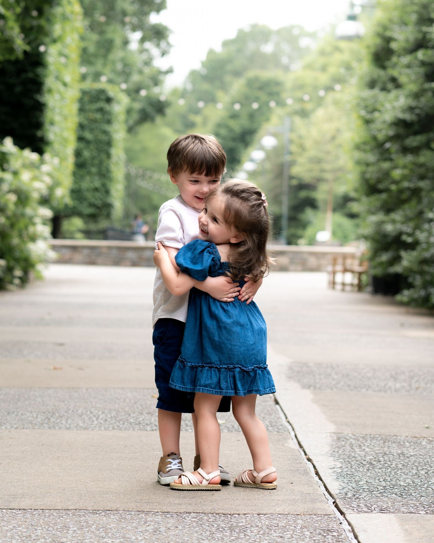 Two young siblings share a sweet hug on a garden pathway surrounded by lush green hedges.
