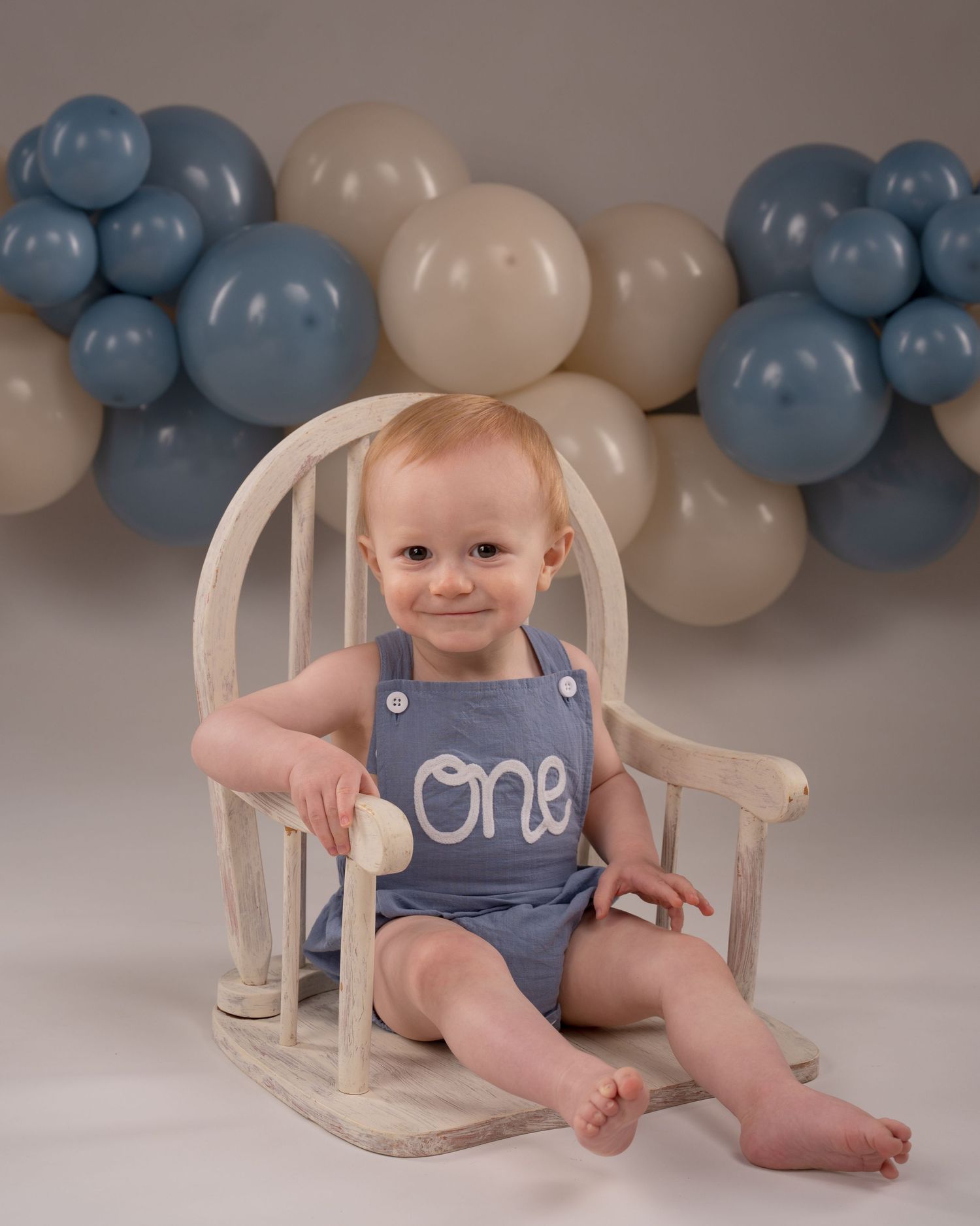 Happy redhead baby boy in blue romper sits in wooden chair with blue and beige balloon garland.