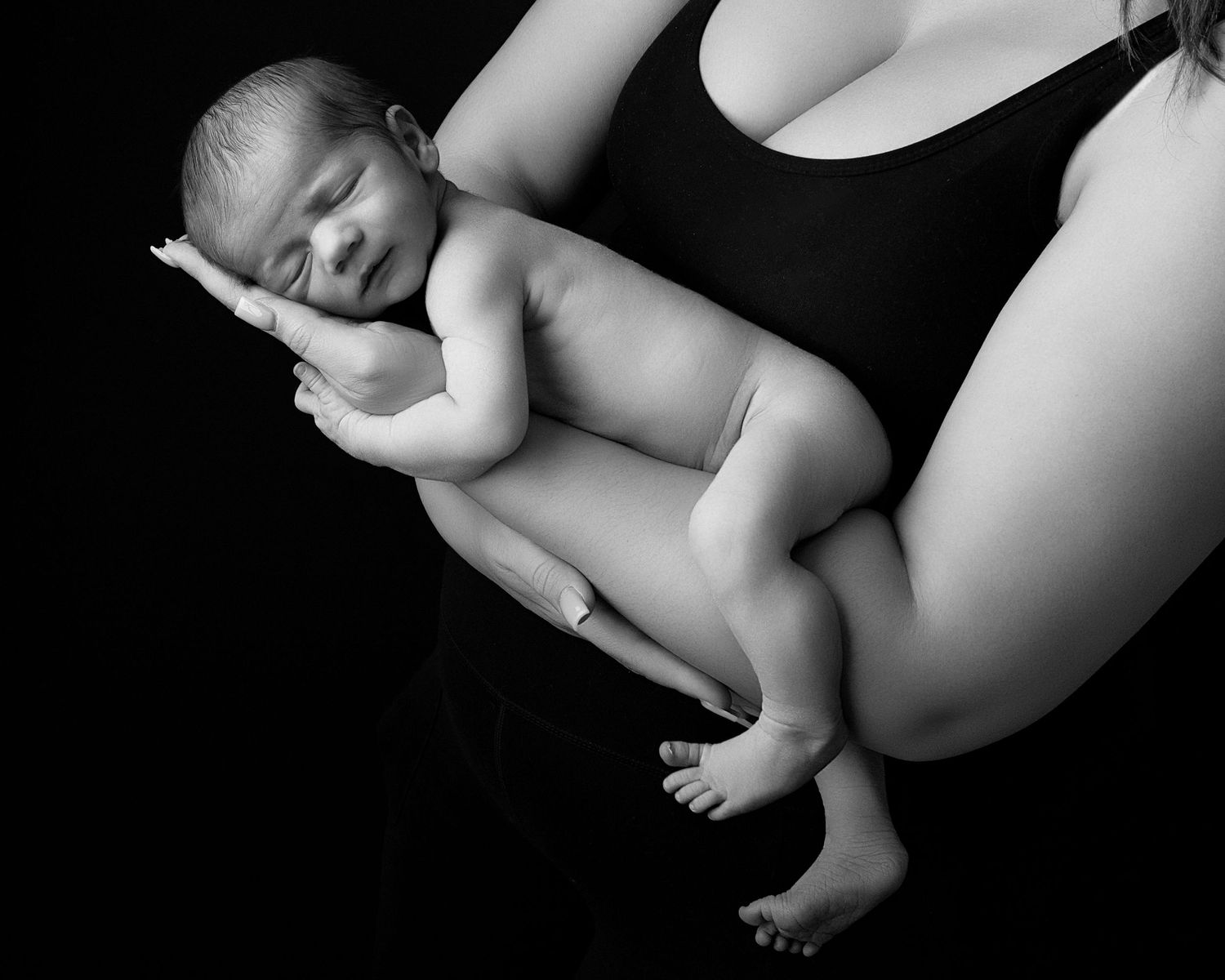 Black and white photo of newborn baby resting on mothers hip against dark background.