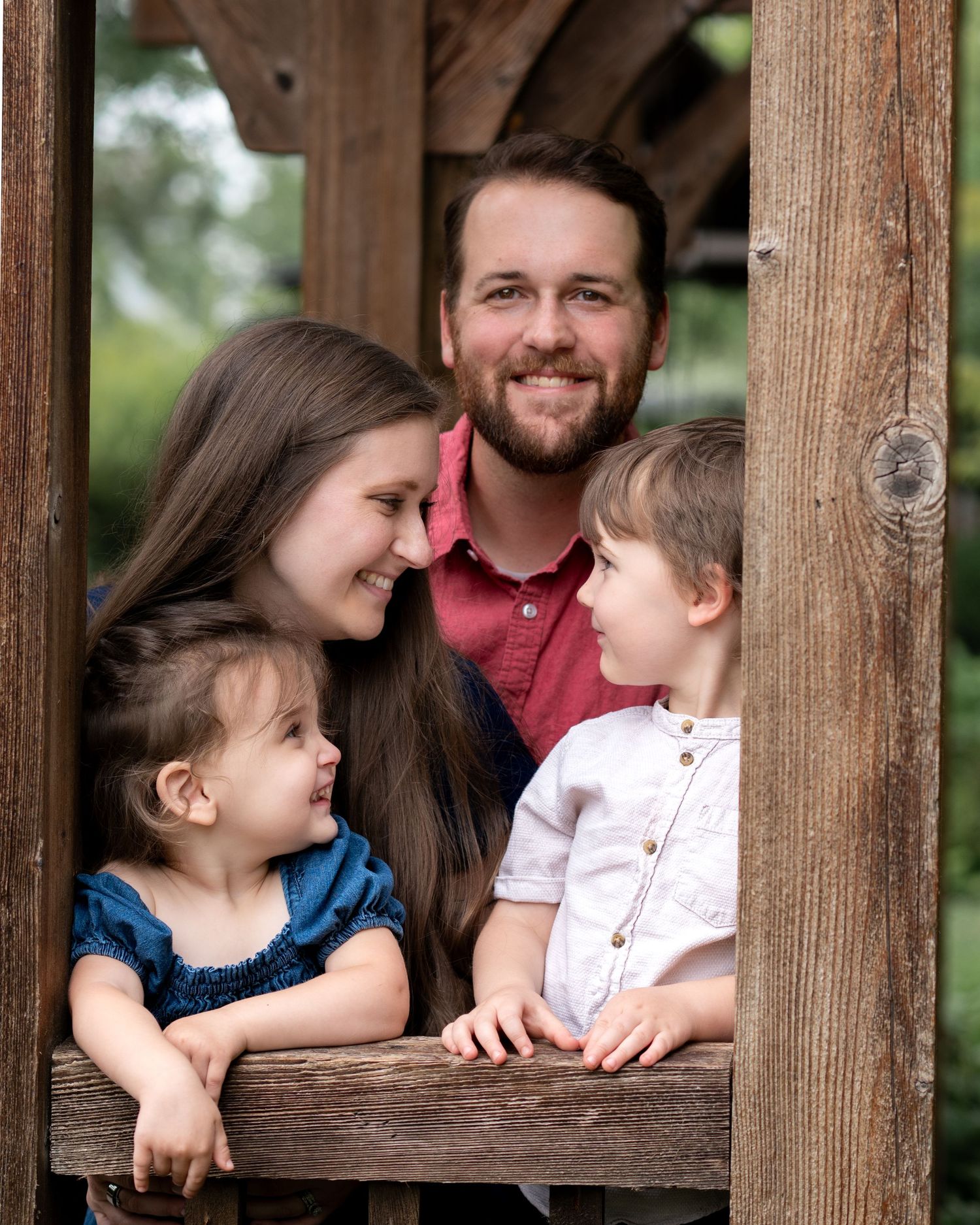 Family of four peeking through a rustic wooden window frame, smiling and laughing together.