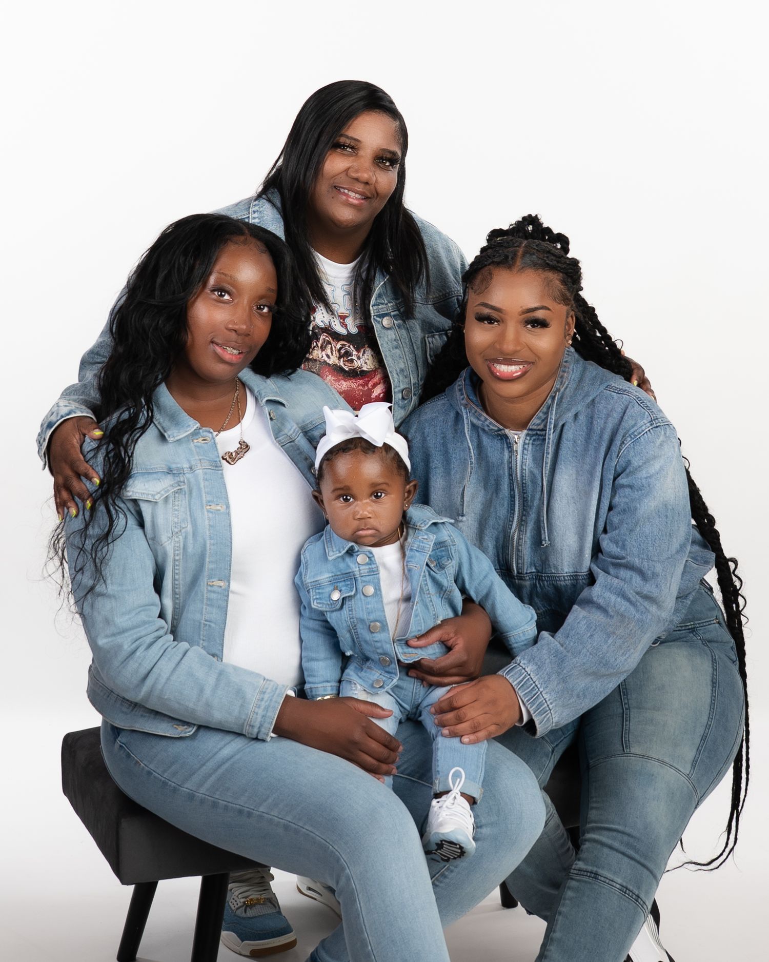 Three generations of women in denim outfits pose together with baby girl in studio portrait.