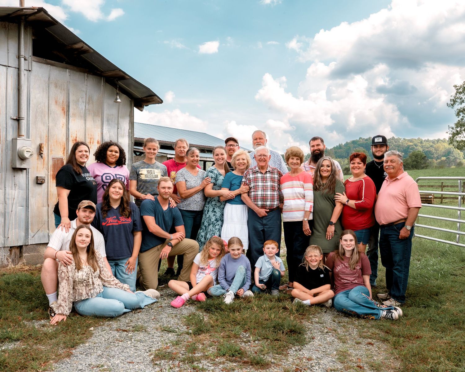 Large multigenerational family group portrait outdoors on a farm with rolling hills and cloudy sky backdrop.