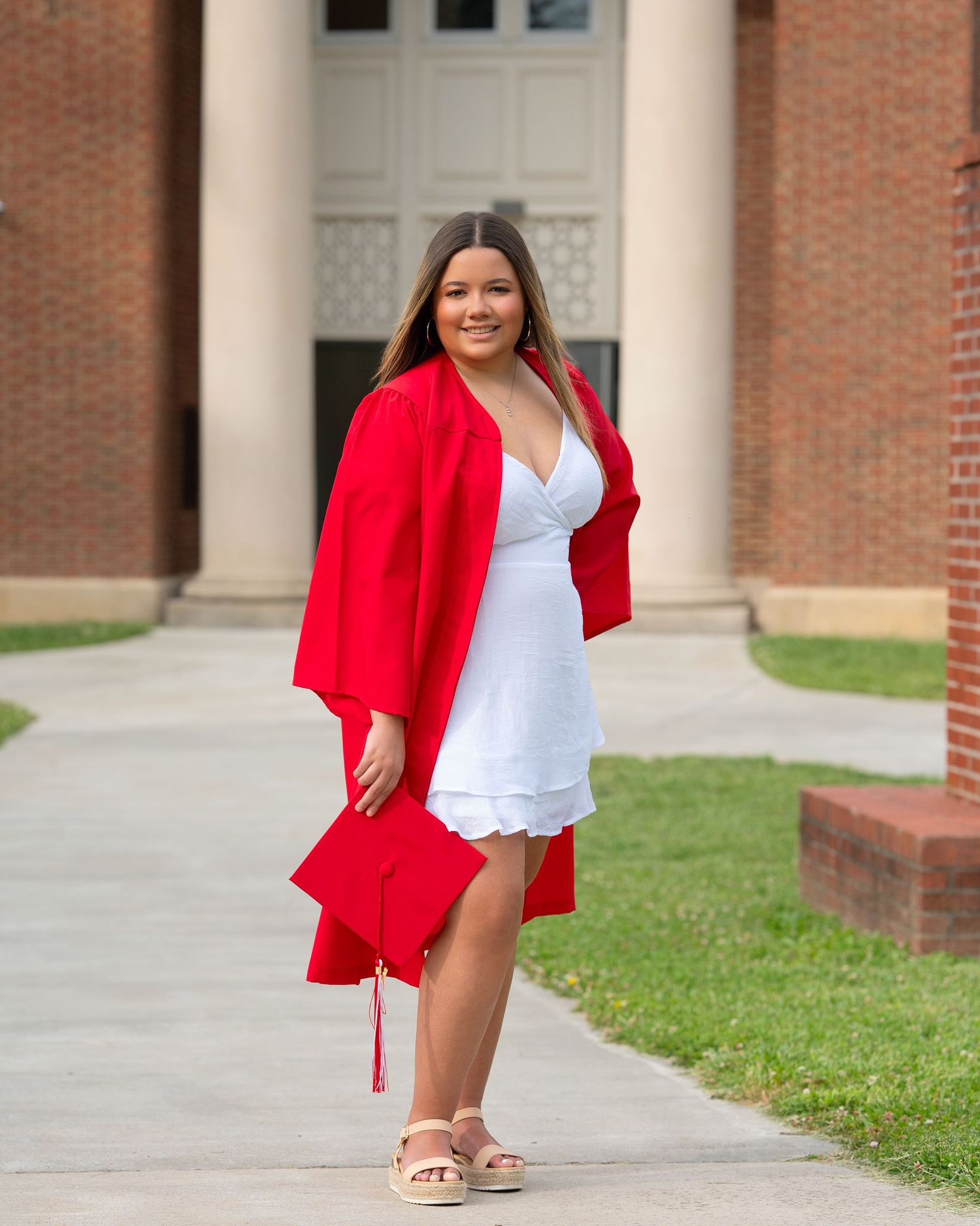 Smiling graduate poses in red cap and gown over white dress outside brick building.