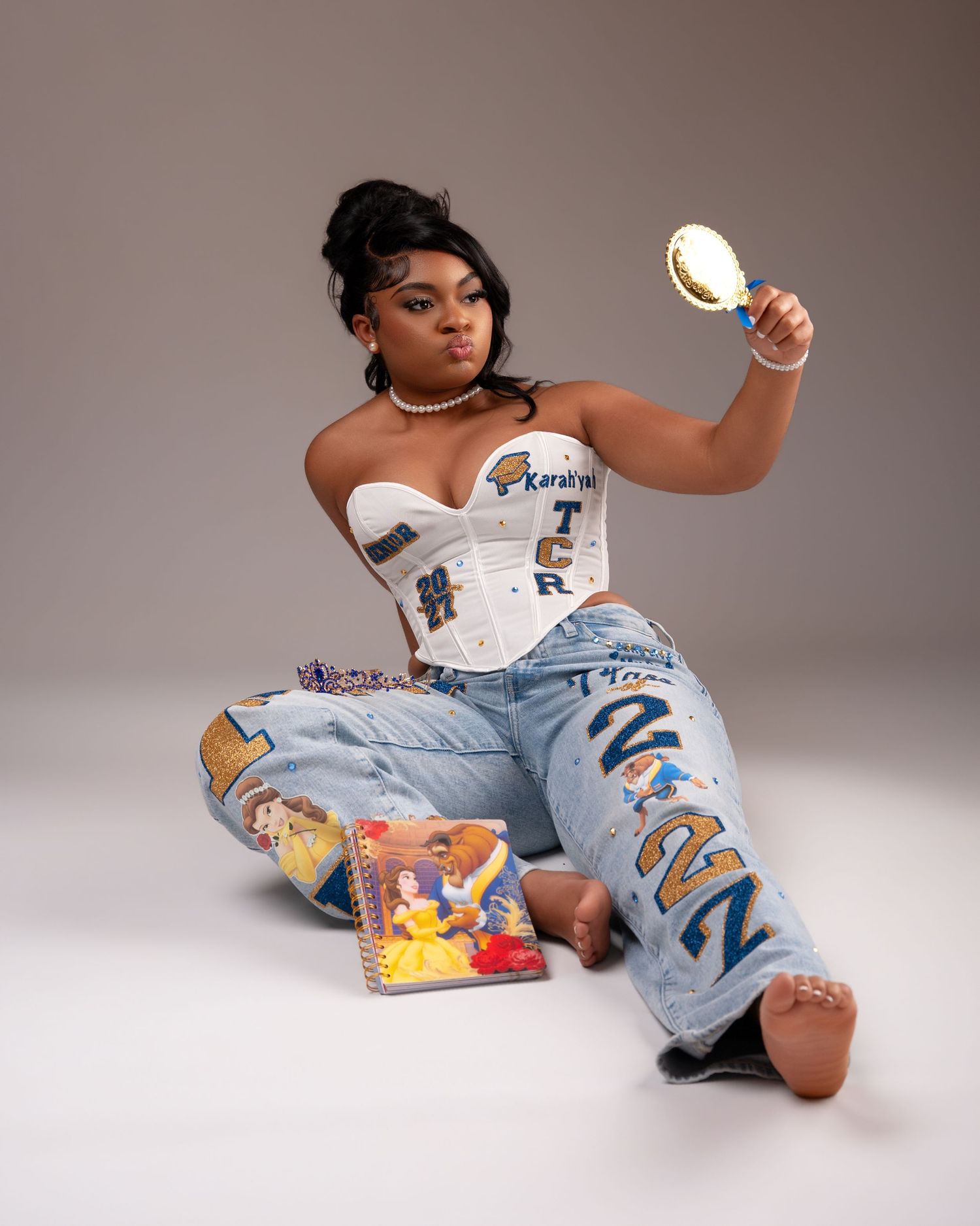 Young woman in white corset top and custom senior jeans holds mirror while sitting on studio floor.