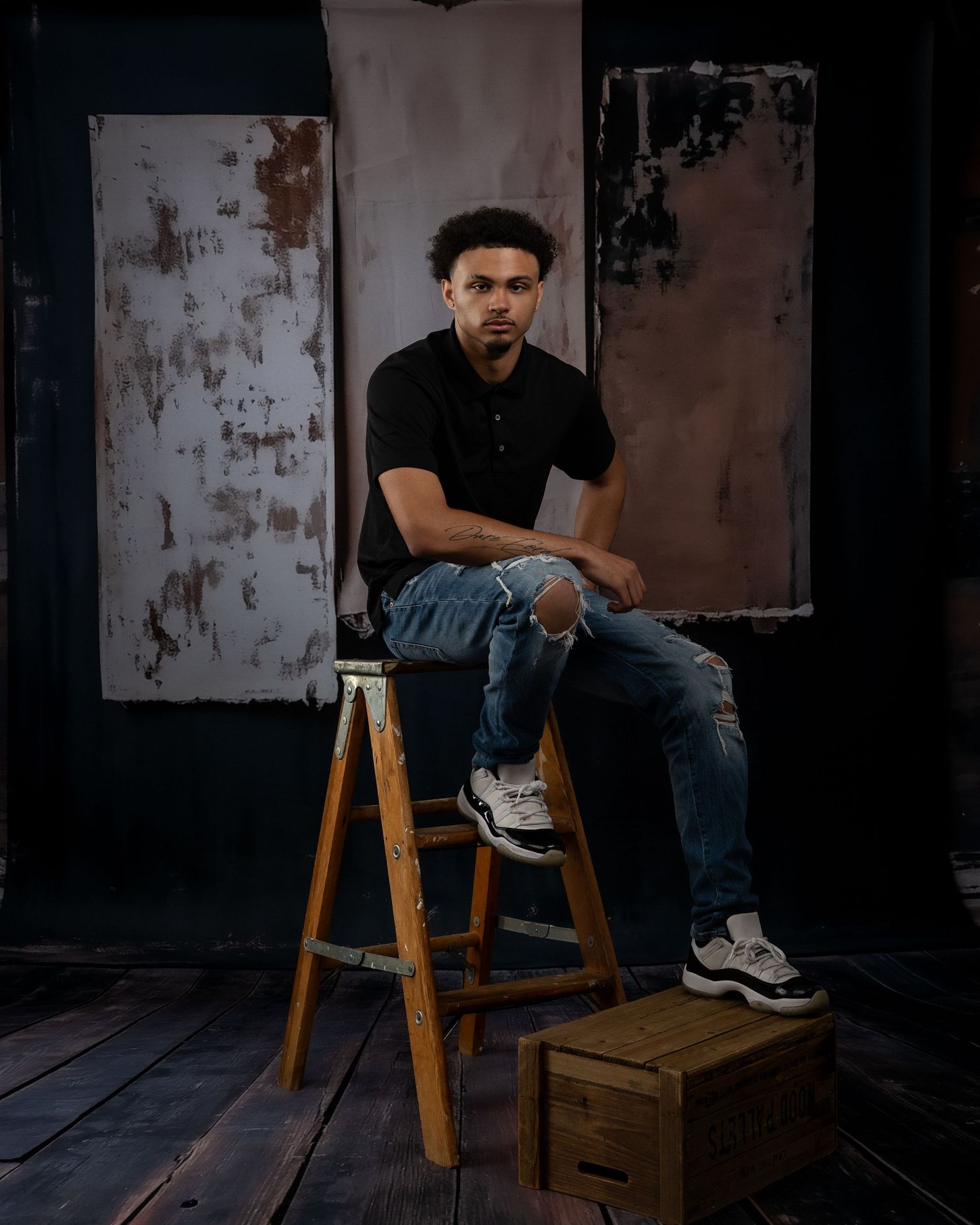 Young man in black polo and ripped jeans sits on wooden ladder stool in artistic studio setting.