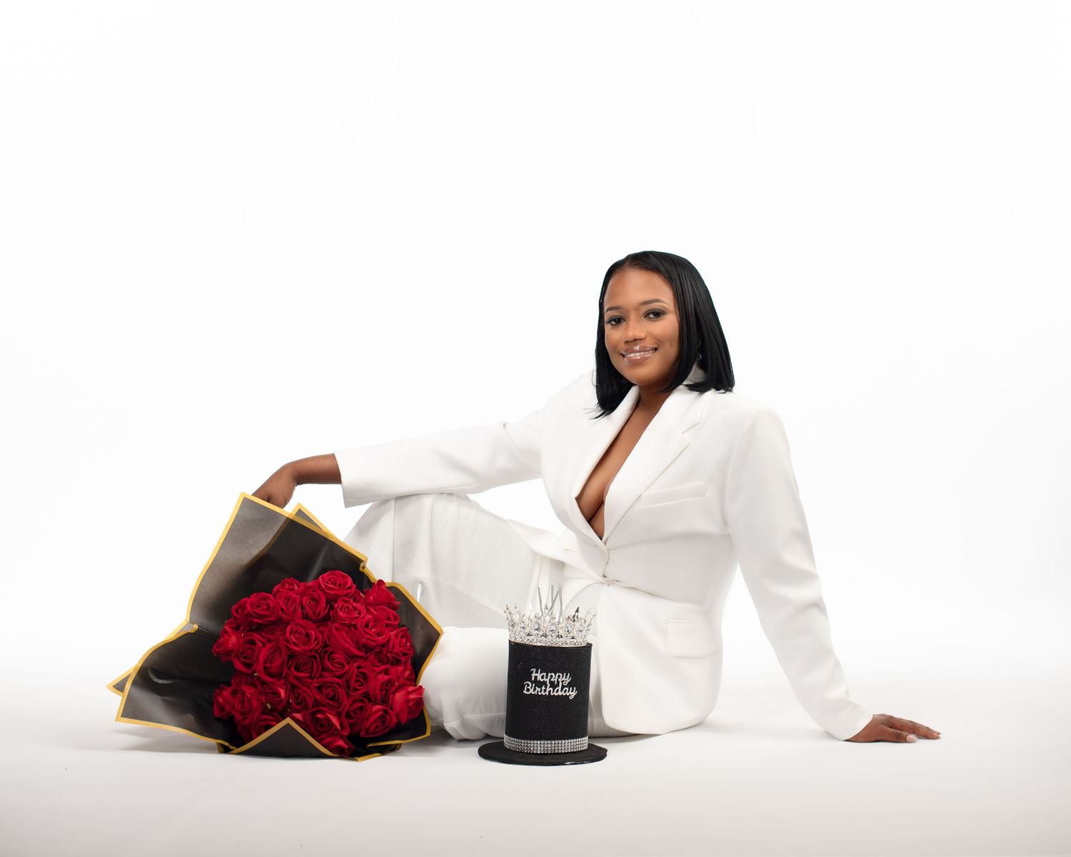 Woman in a white blazer and pants sits on a white floor beside a bouquet of red roses and a black Happy Birthday cake with a crown topper.