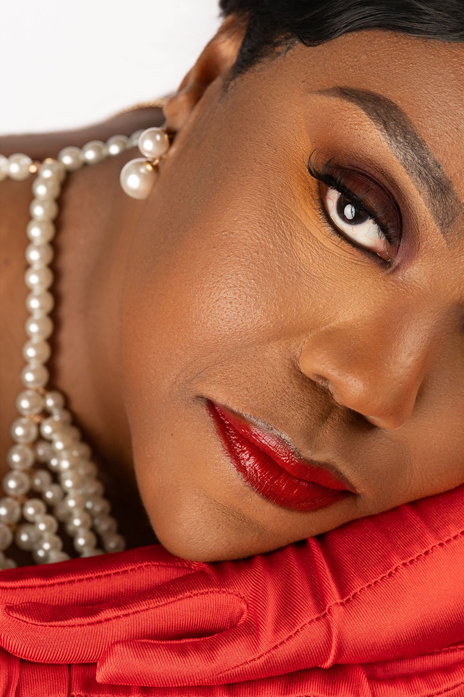 Close-up beauty portrait of a woman with bold red lipstick, bronze eyeshadow, pearl necklace and earrings, resting on a red satin glove.
