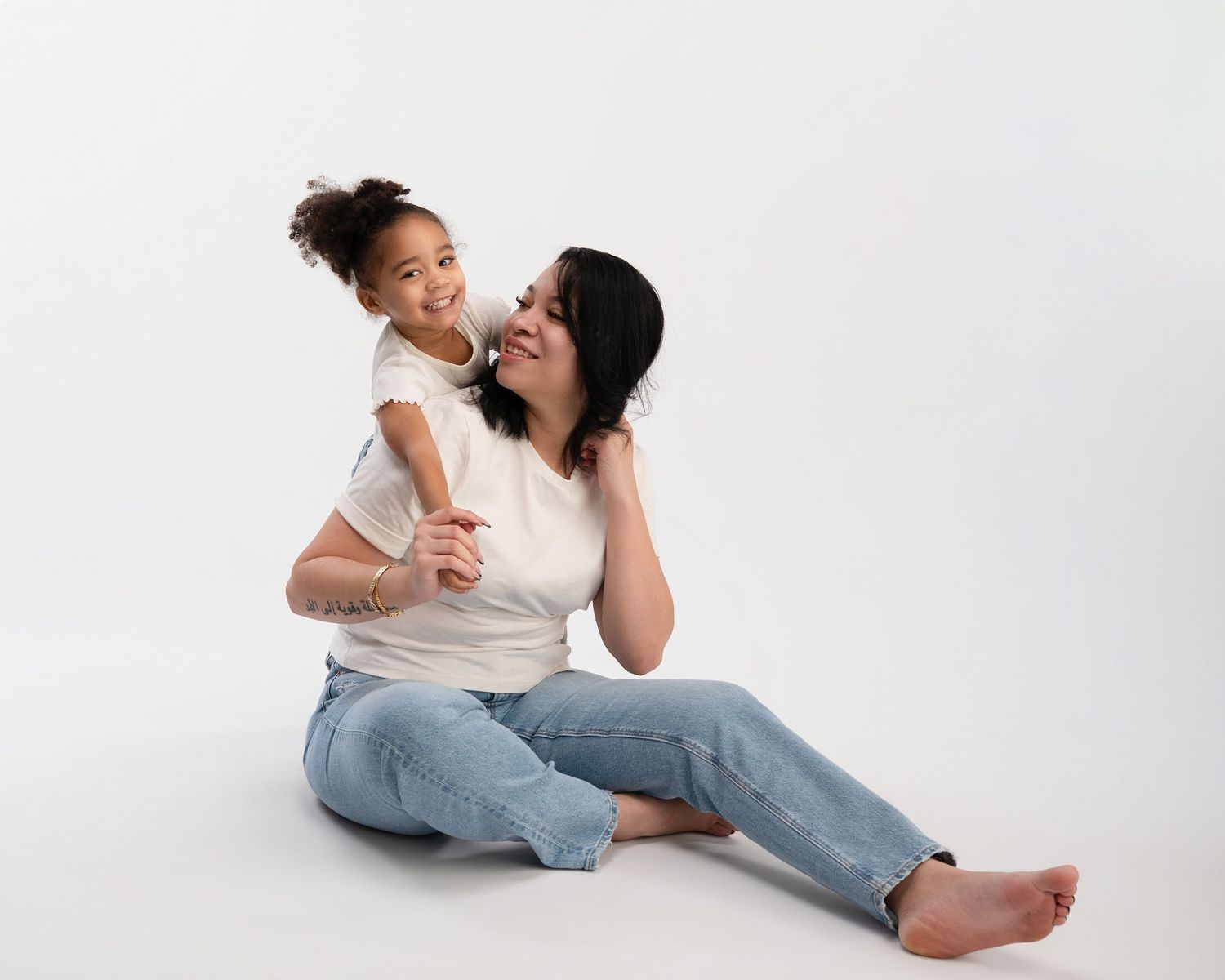 Mother sits on floor smiling as young daughter hugs her from behind in white studio.