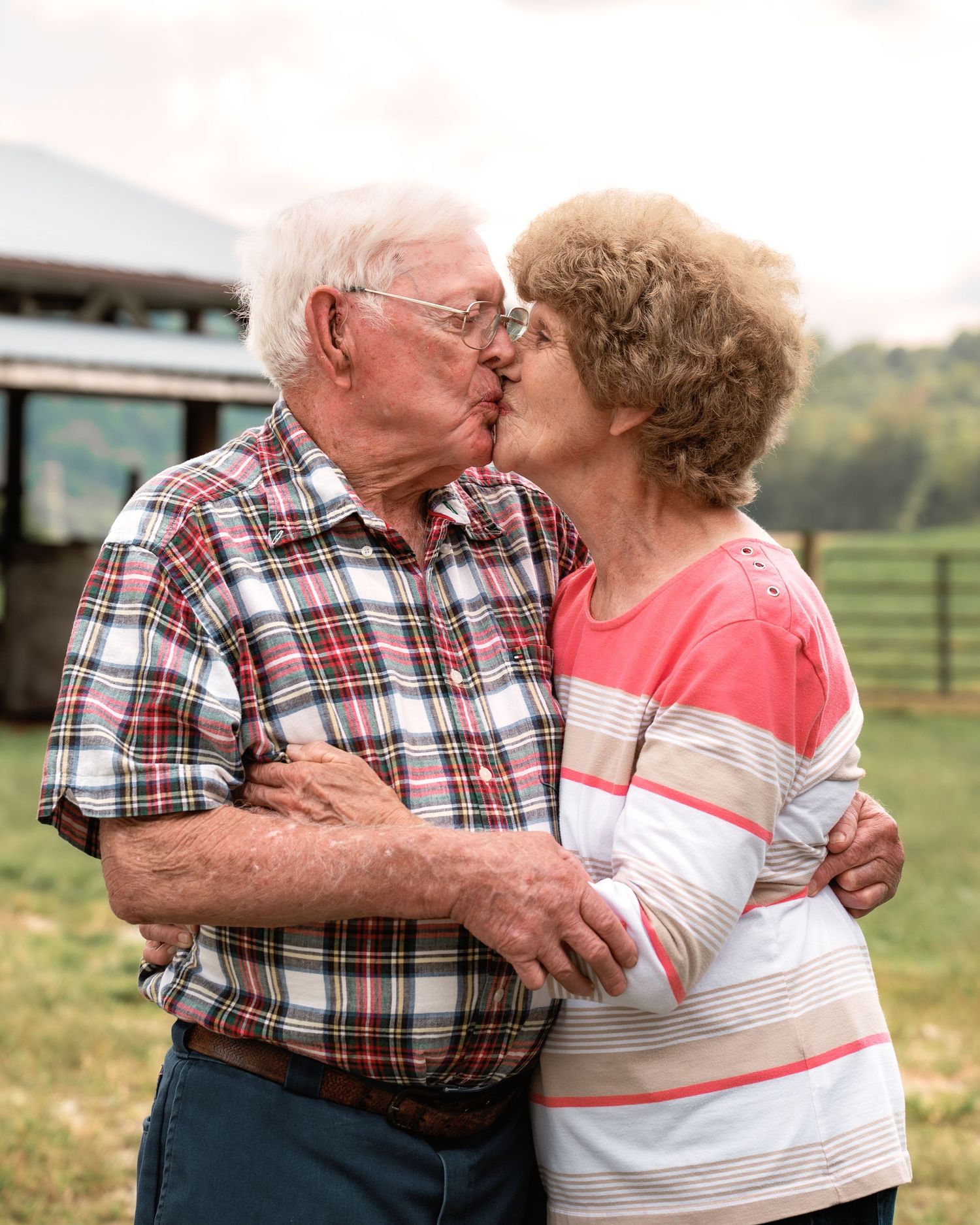 Elderly couple sharing a tender kiss outdoors on a farm with green fields in the background.