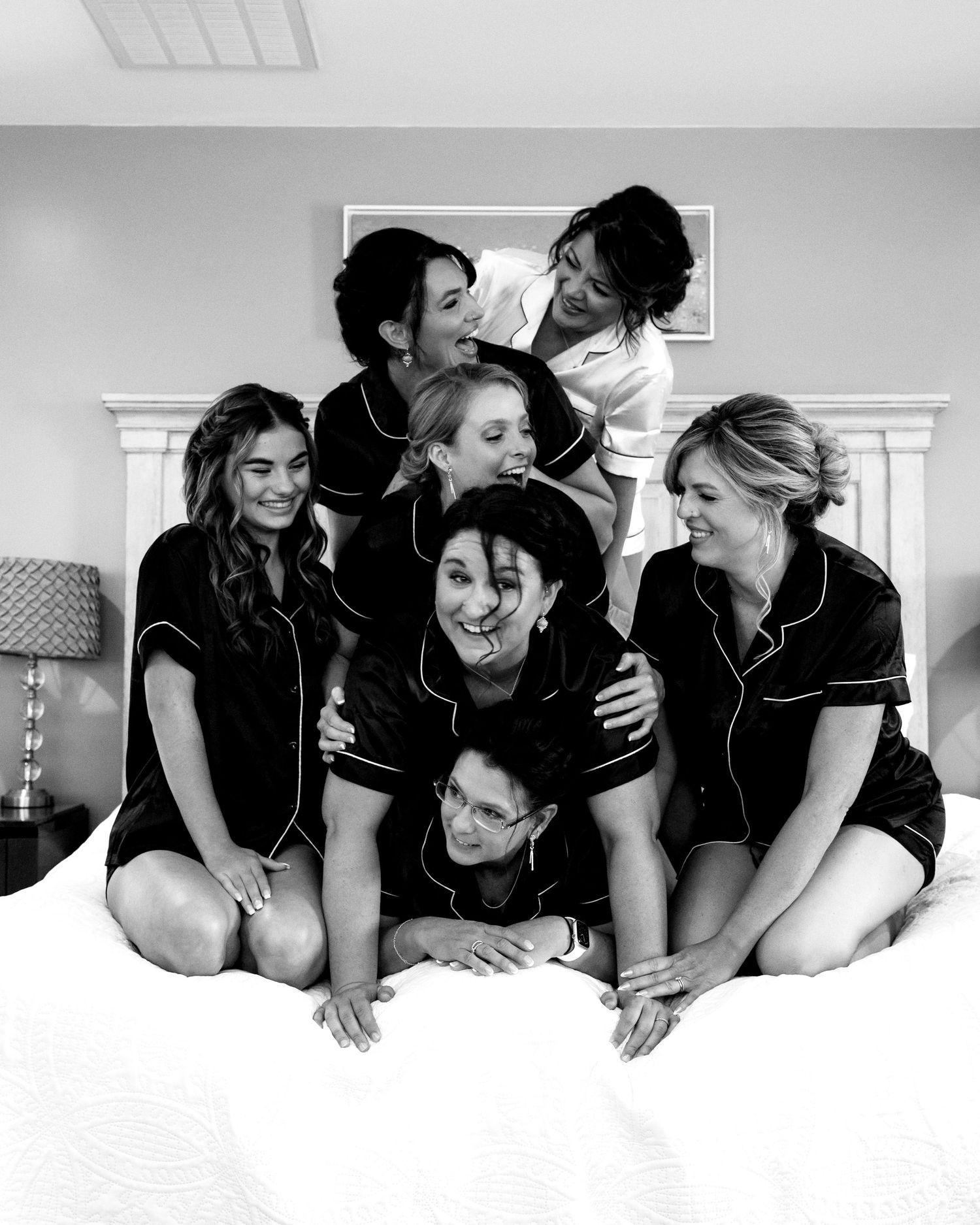 Six bridesmaids in matching black satin pajamas pose playfully on a bed in a black and white photo.