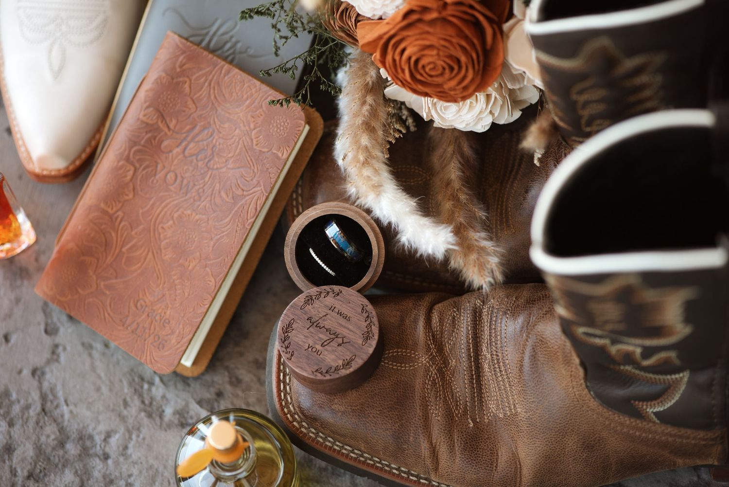 Rustic wedding flat lay featuring cowboy boots, leather journal, ring box, and floral bouquet details.