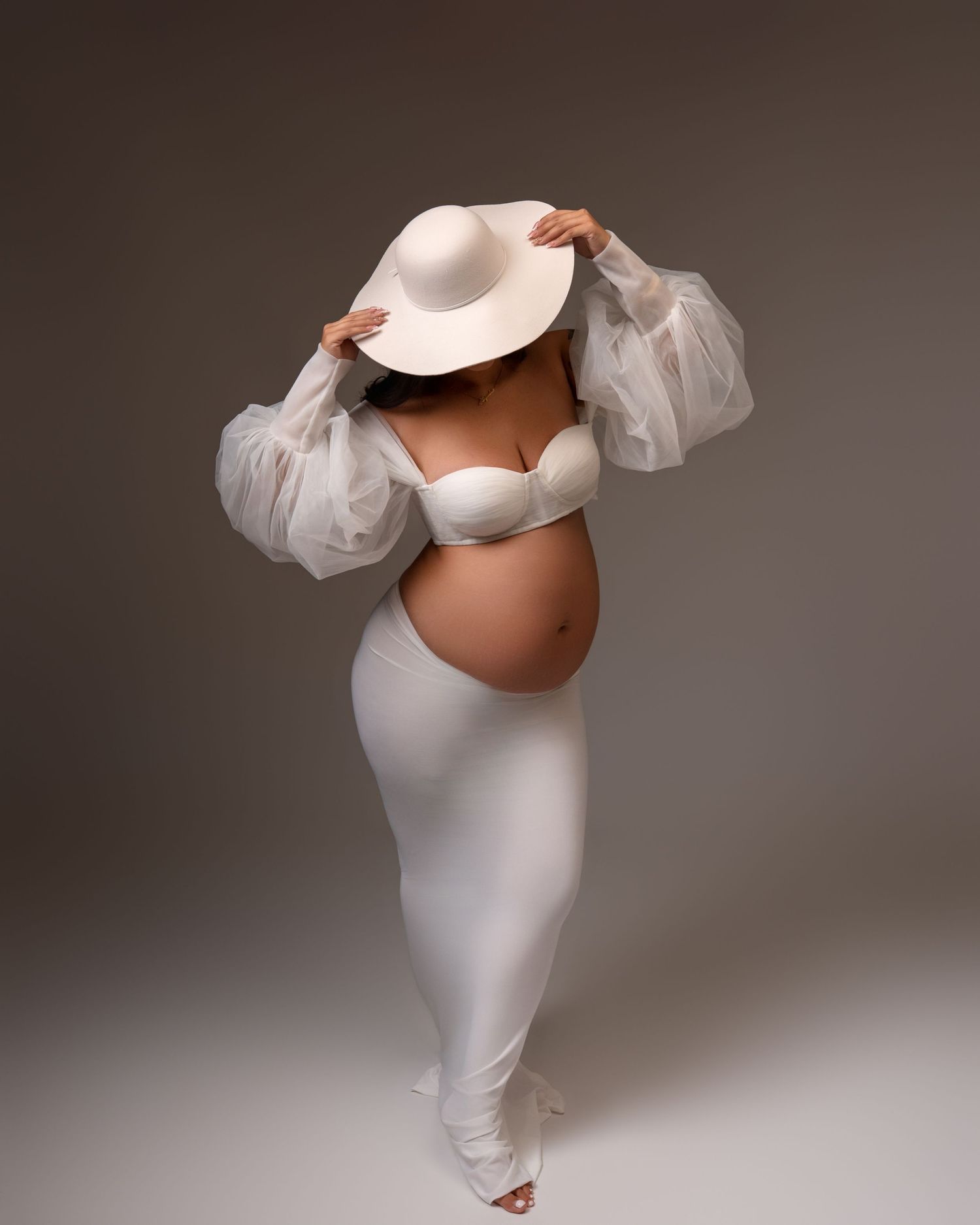 Pregnant woman in white ensemble and wide-brim hat elegantly posing to showcase baby bump in studio.