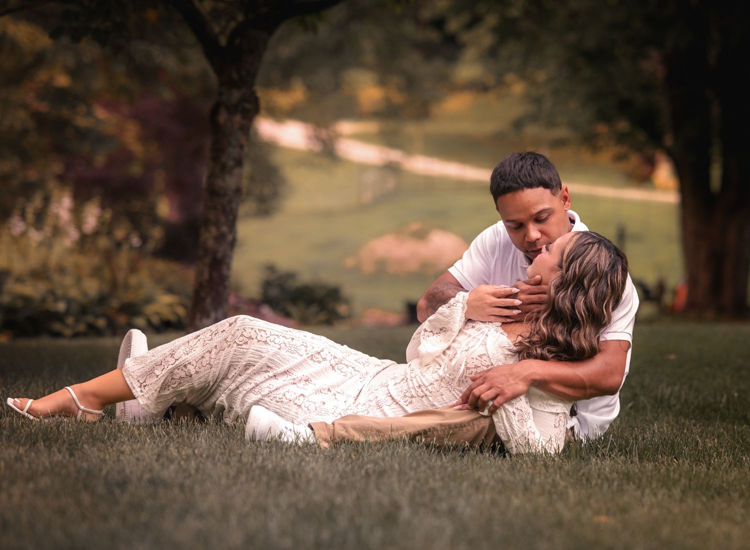 Couple sharing an intimate moment on grass, man leaning over woman in white lace dress outdoors.