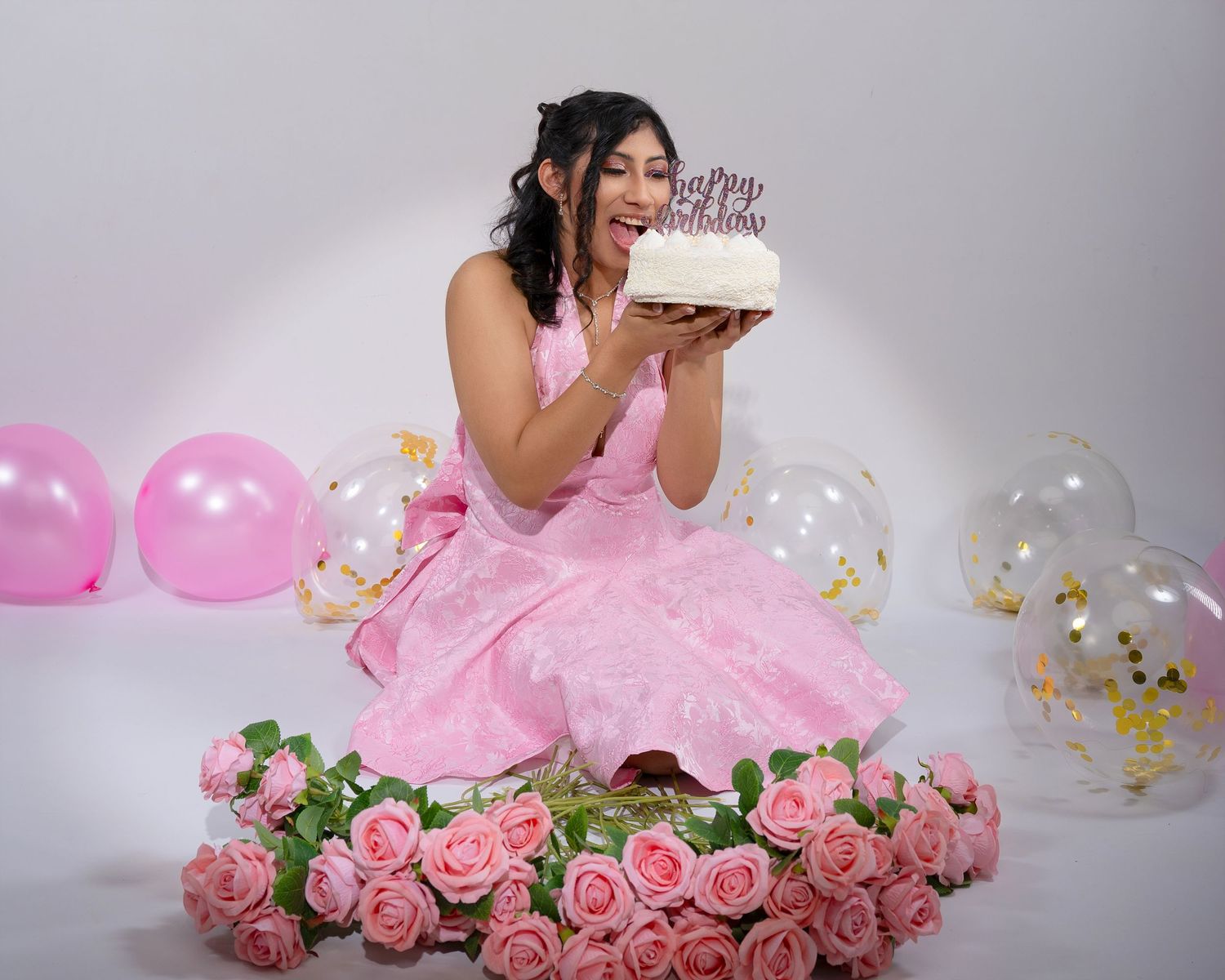 Young woman in pink dress celebrating birthday with cake, balloons, and roses in white studio.