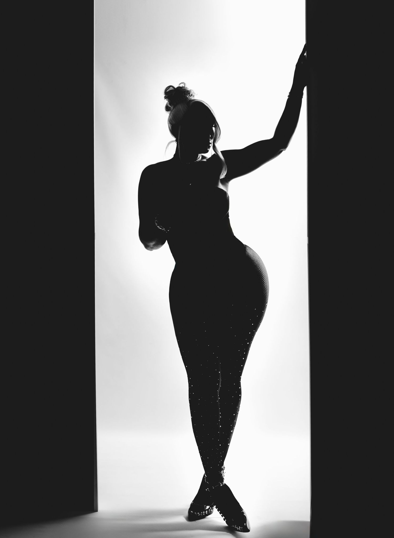 Striking black and white silhouette of curvy woman in sequined catsuit posing in doorway with dramatic backlight.