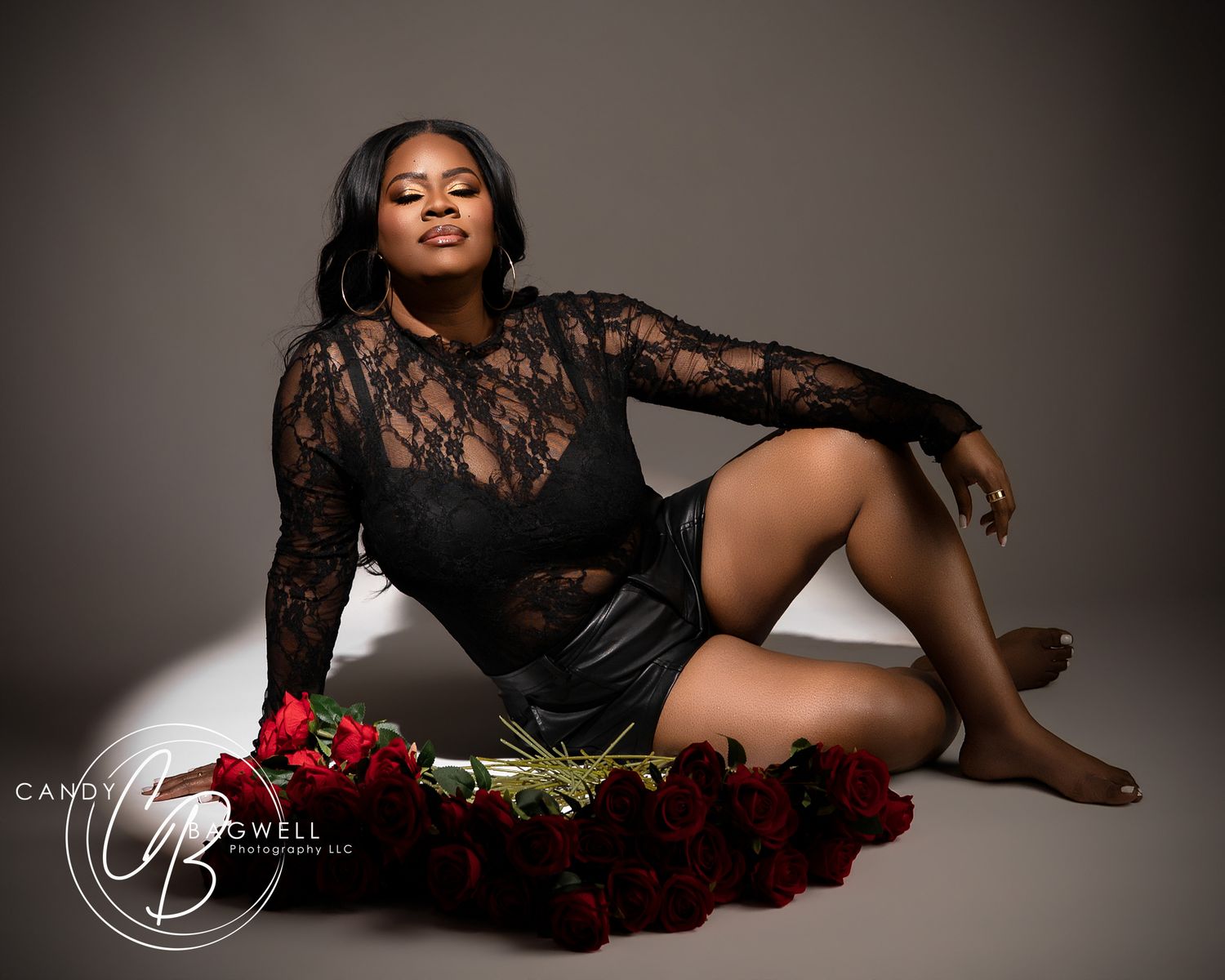 Woman in black lace bodysuit sits beside red roses in studio portrait.
