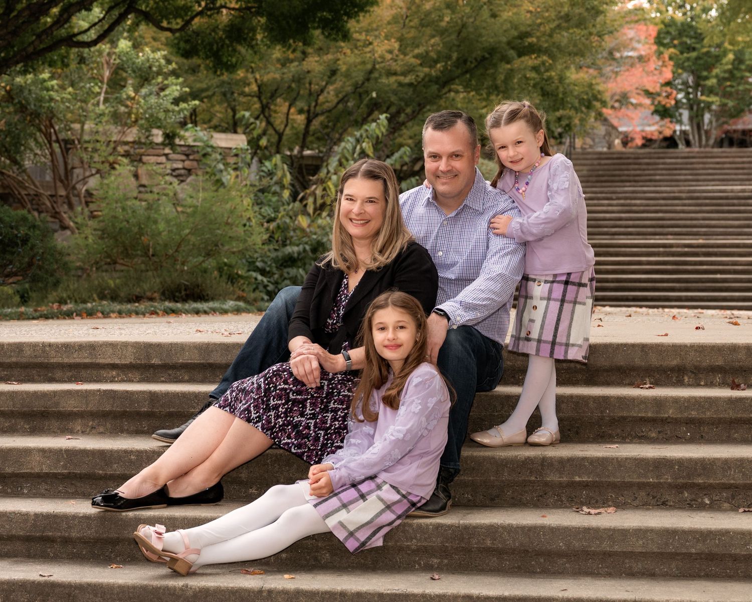 Family of four posing on stone steps surrounded by lush greenery, two girls in purple plaid dresses.