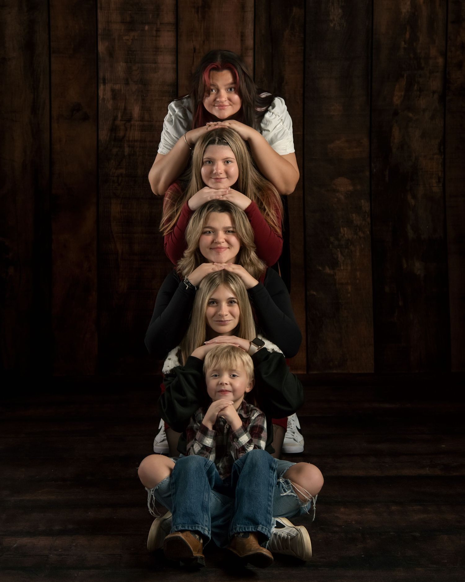 Four siblings pose stacked vertically in fun creative portrait against rustic dark wood background.
