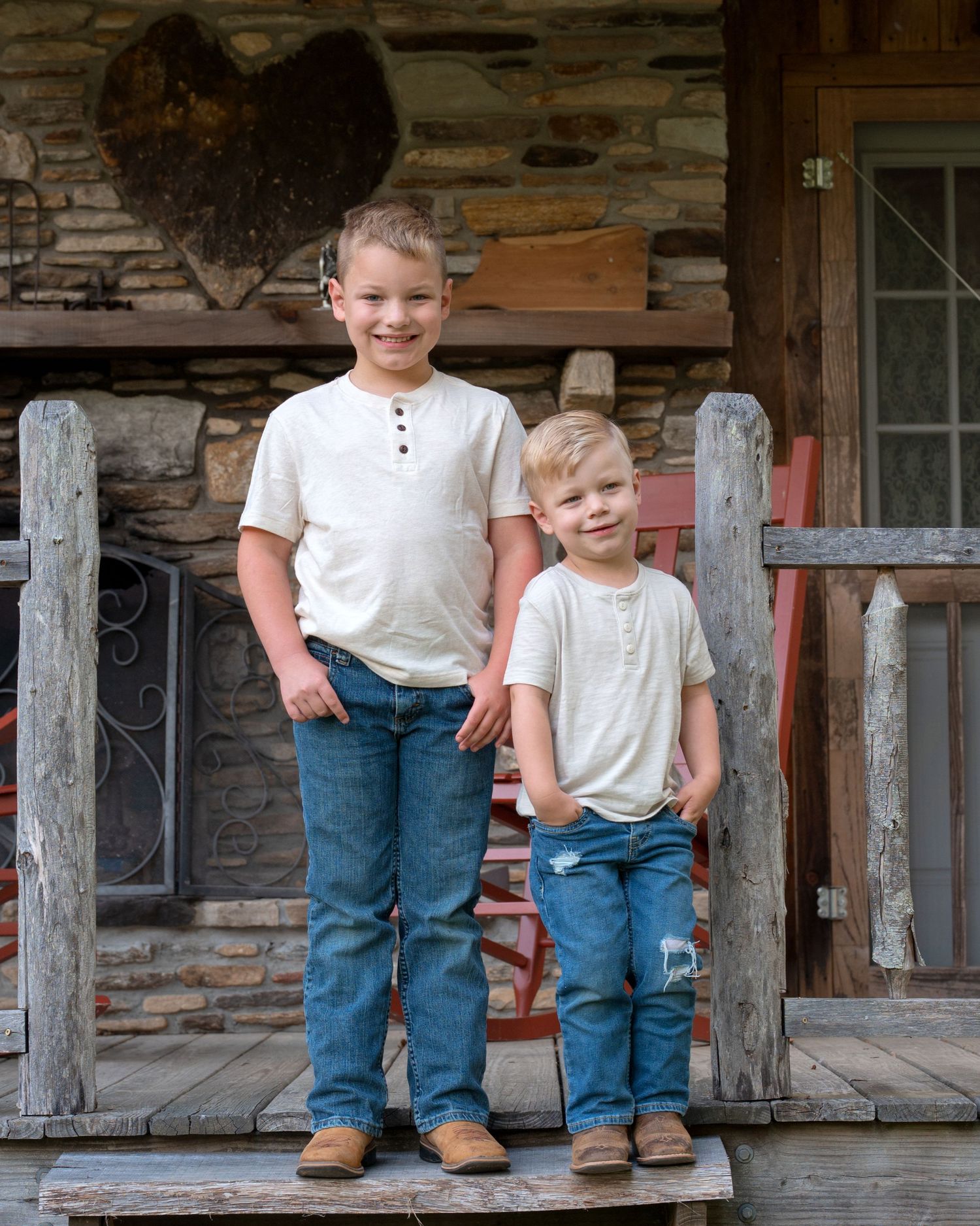 Two young brothers in matching cream henley shirts and jeans pose on a rustic stone porch.
