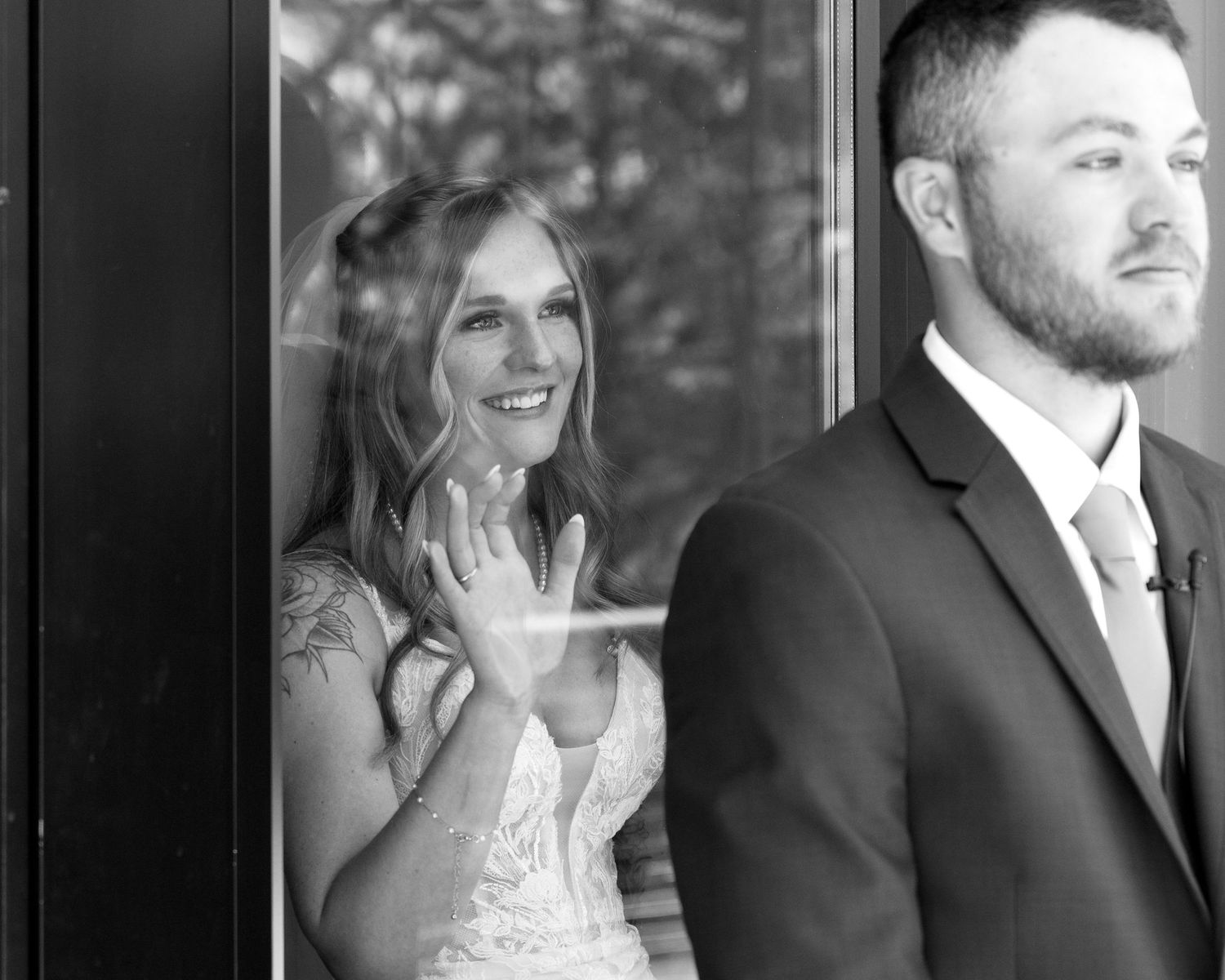 Black and white photo of bride waving through glass door while bearded groom stands outside in suit.