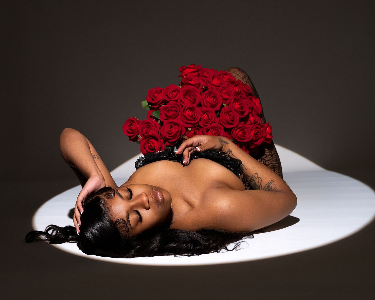 Woman with long dark hair lying on a white circular backdrop, resting her head on her hand and holding a bouquet of red roses.