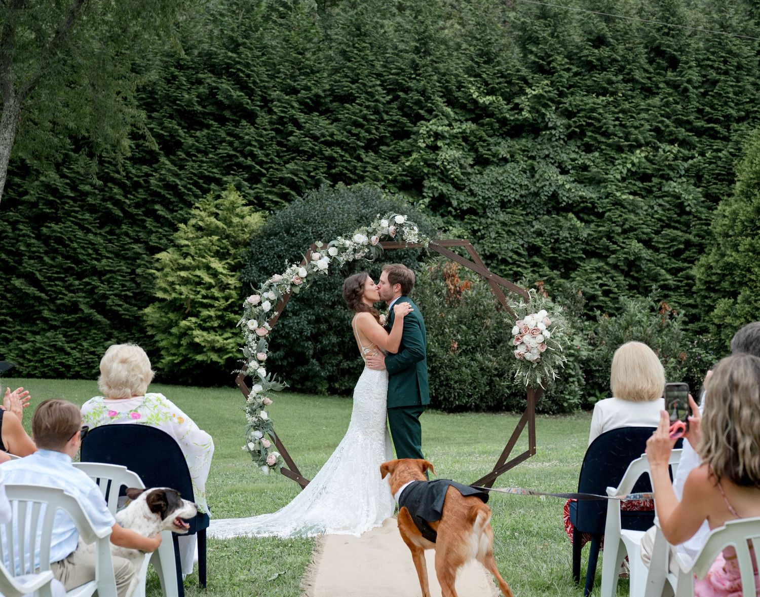 Bride and groom share first kiss at outdoor garden wedding ceremony with floral arch.