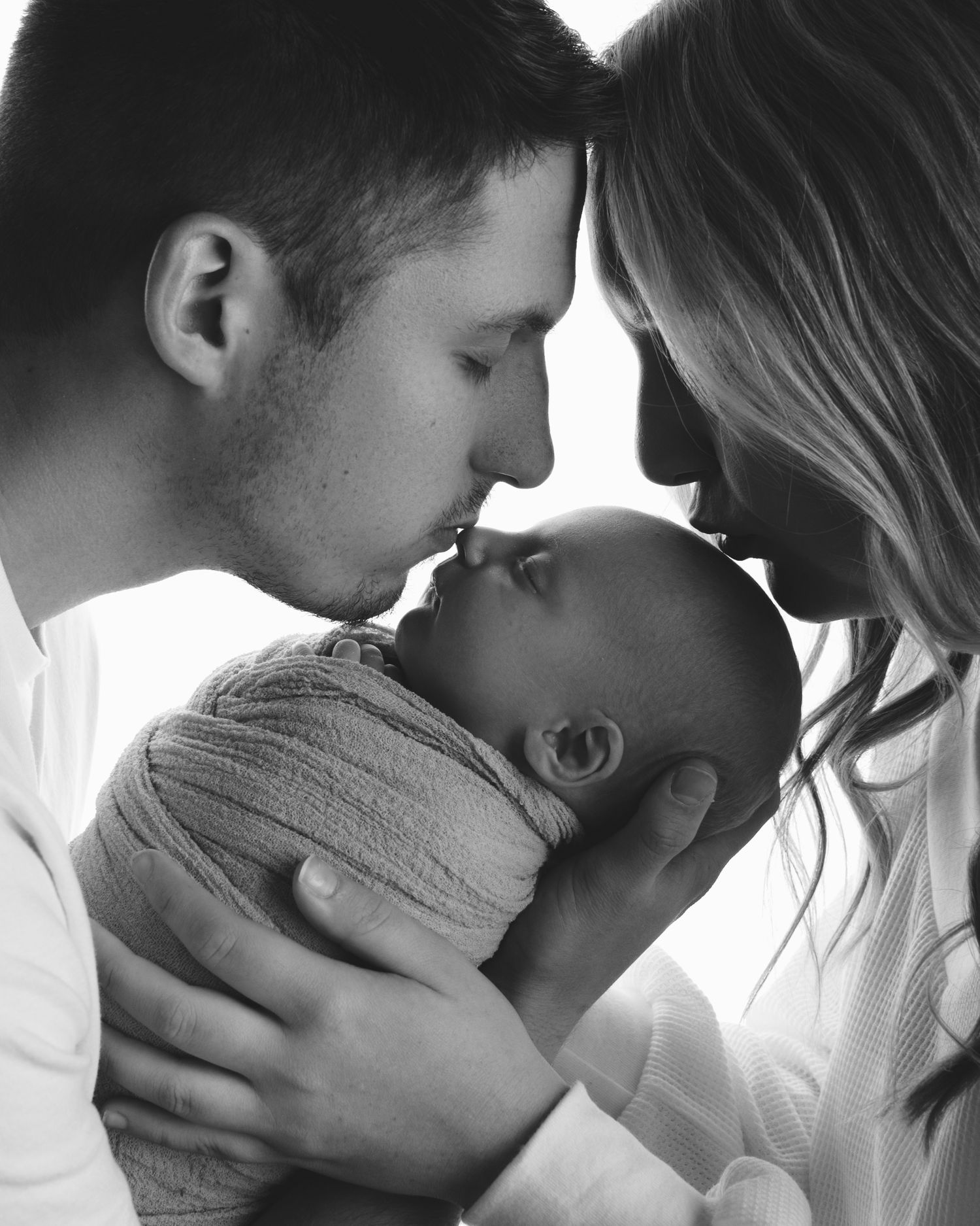 Black and white portrait of new parents leaning in to kiss their swaddled newborn baby together.